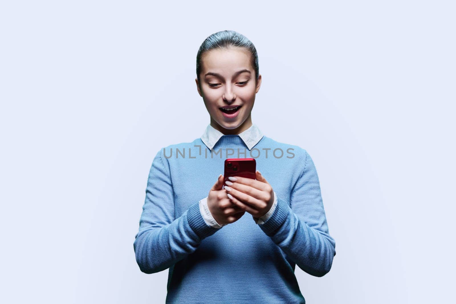 Surprised teenage student, girl 15, 16 years old using smartphone on white studio background. Adolescence, internet technologies, mobile online applications apps services for learning leisure