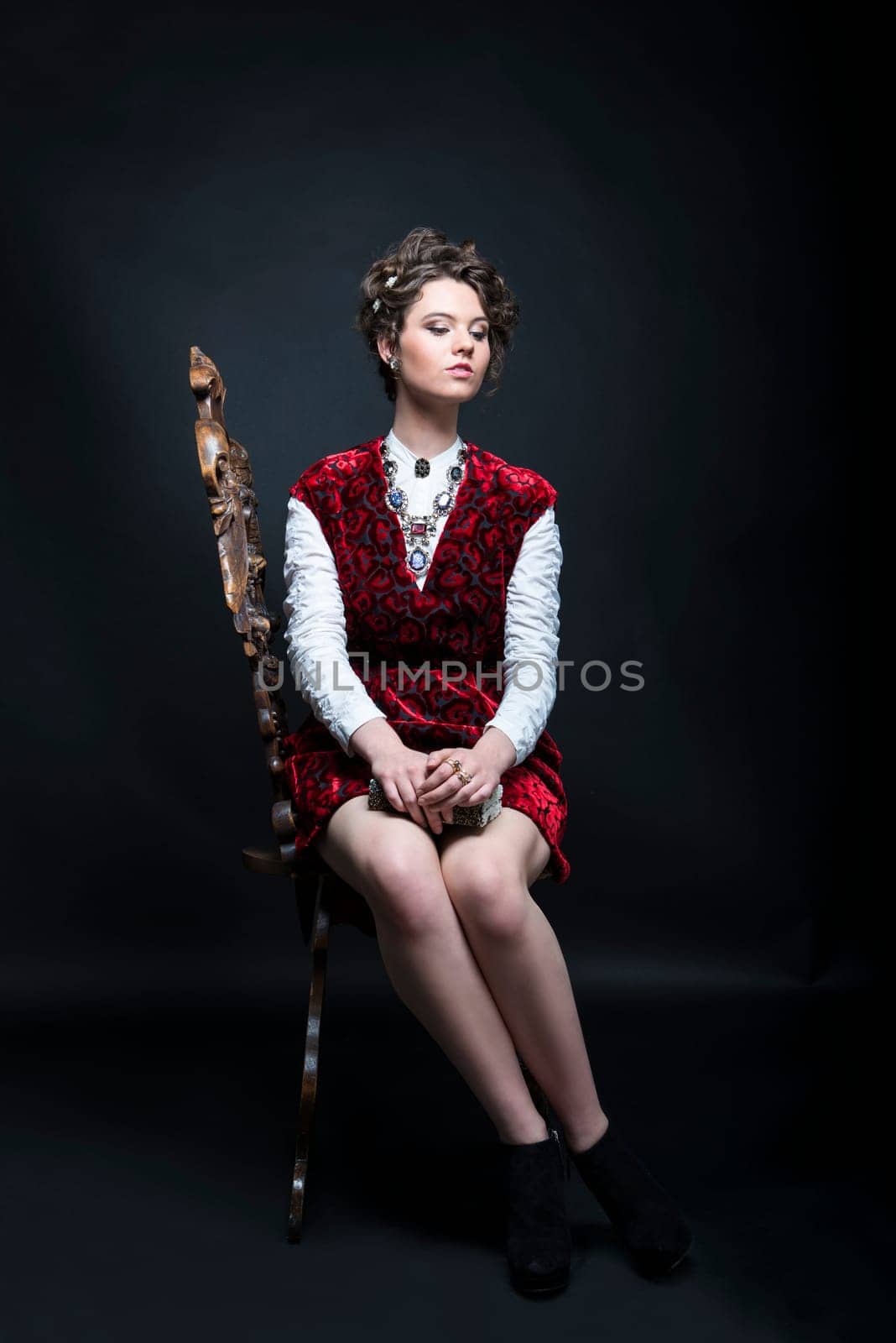 Portrait of beautiful Caucasian model in red Victorian-style velvet dress, seated on wooden chair