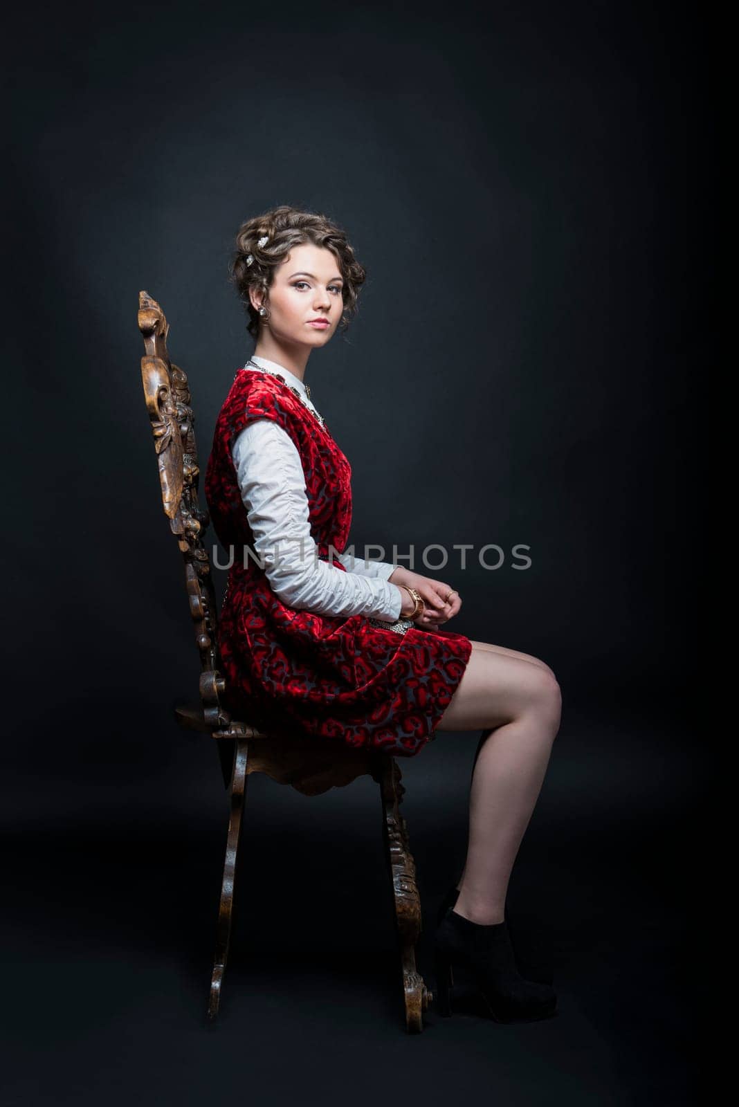 Portrait of beautiful Caucasian model in red Victorian-style velvet dress, seated on wooden chair