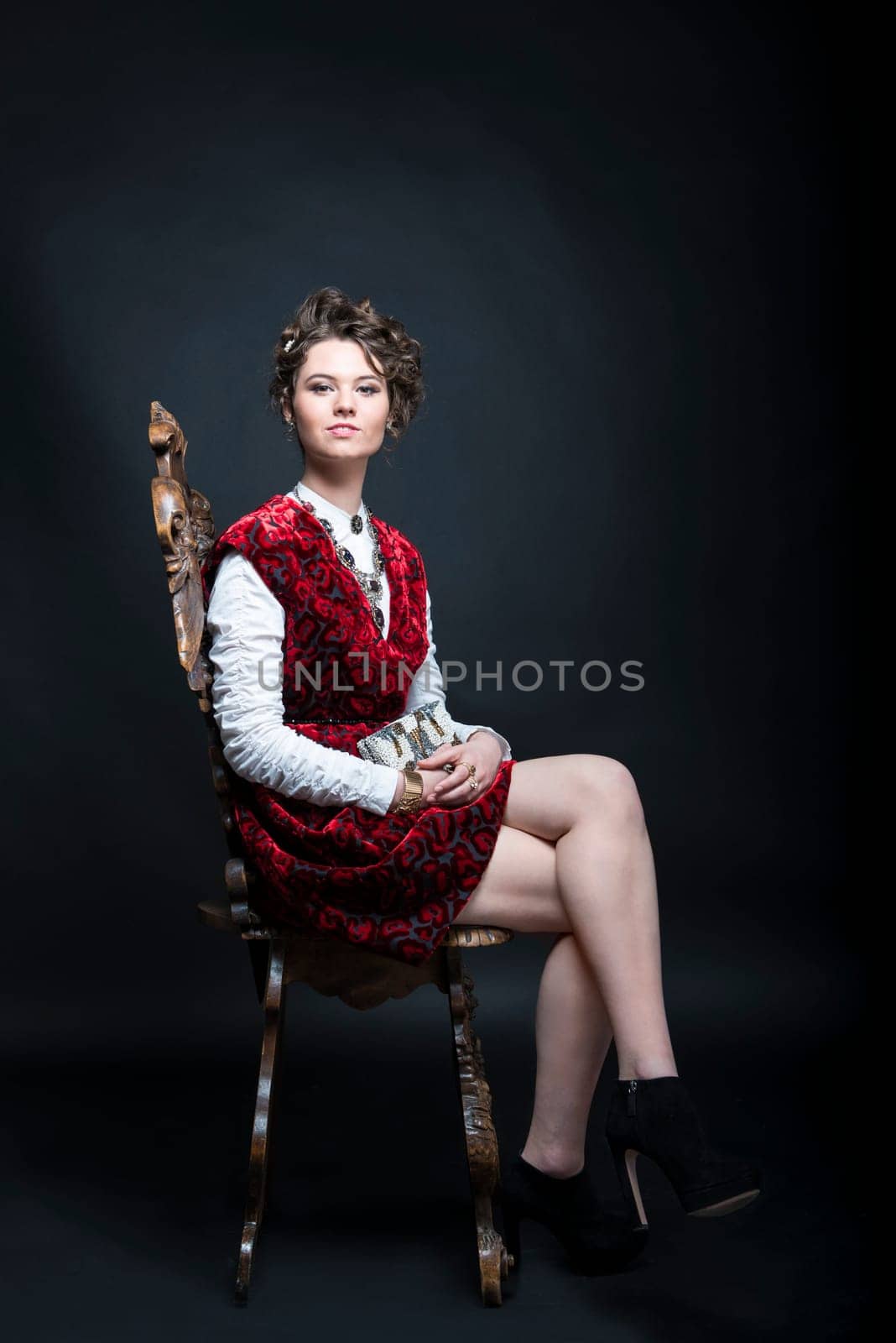 Portrait of beautiful Caucasian model in red Victorian-style velvet dress, seated on wooden chair
