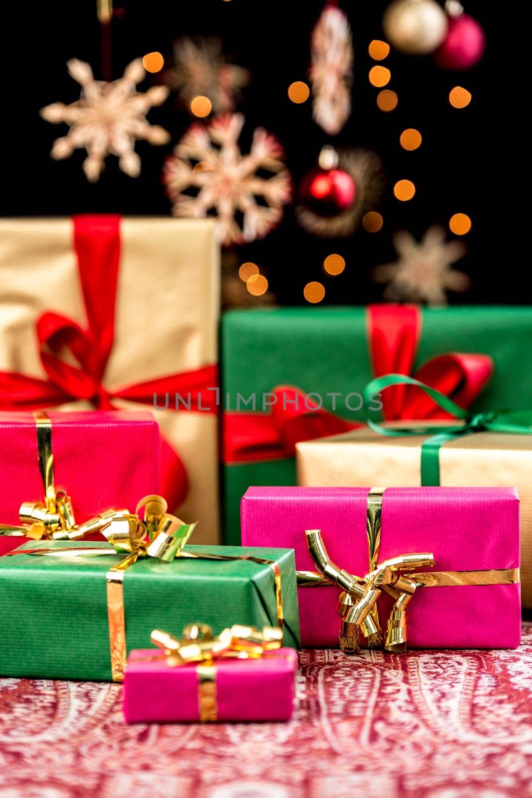 Christmas gifts in red,green and gold on a festive blanket. The focus is on the larger magenta-coloured package. The orange candlelight reflections sparkle between the blurry Christmas tree domes and straw stars. Christmas gifts in red,green and gold placed on a festive cloth. Focus is on the bigger magenta present. Orange candle light twinkles do intertwine with blurred baubles and stars in the back.