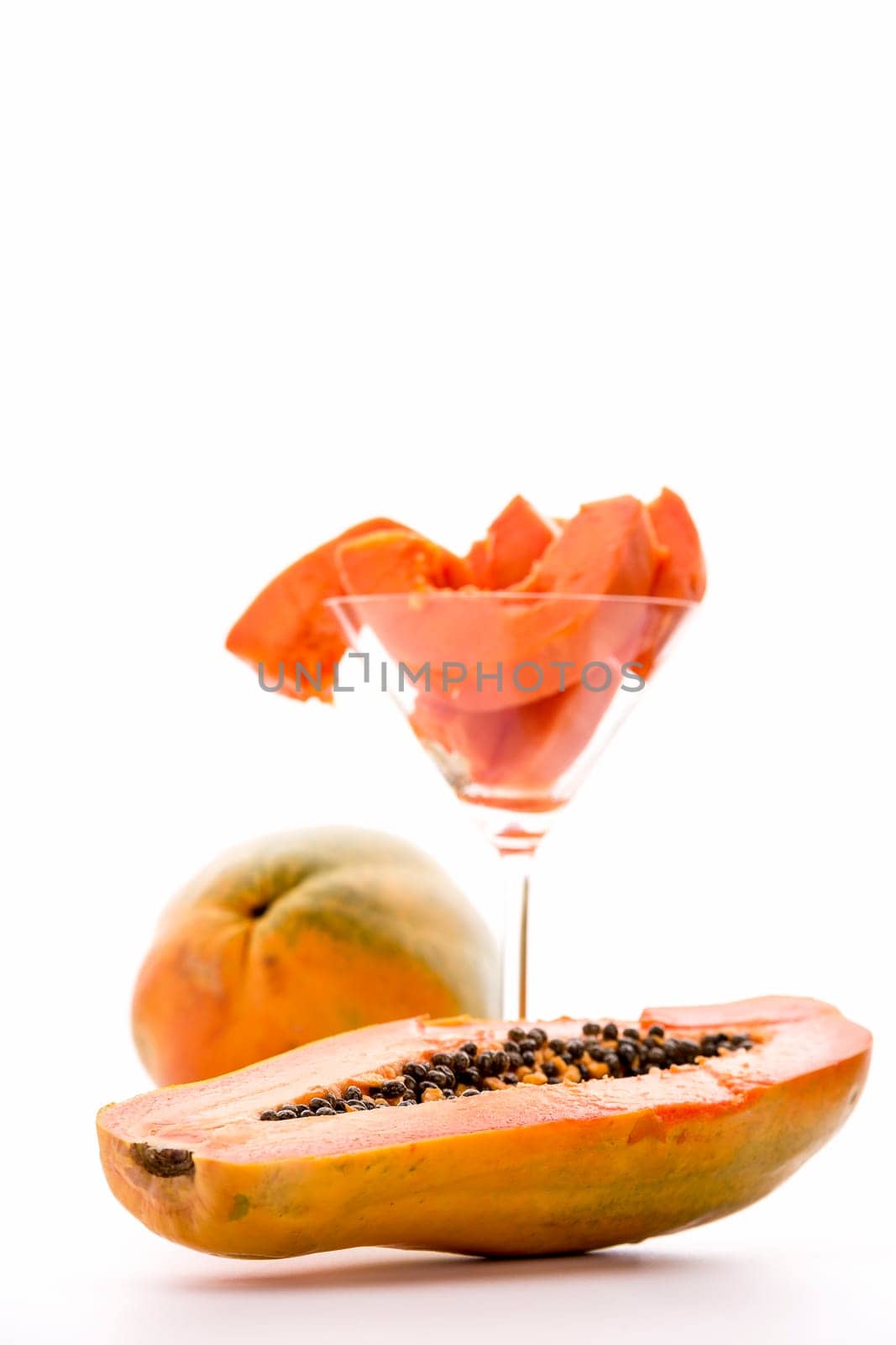 A halved pawpaw fruit in focus in the foreground. In the background is an entire fruit and a glass filled with its fruit pulp..
