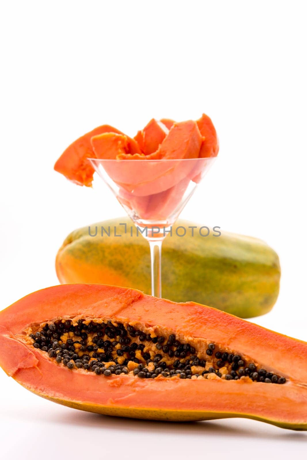 A longitudinal section through the globose body of a papaya fruit. Behind it,there is a cocktail glass filled with peeled papaya slices and an entire fruit. The focus is on the halved fruit in front..