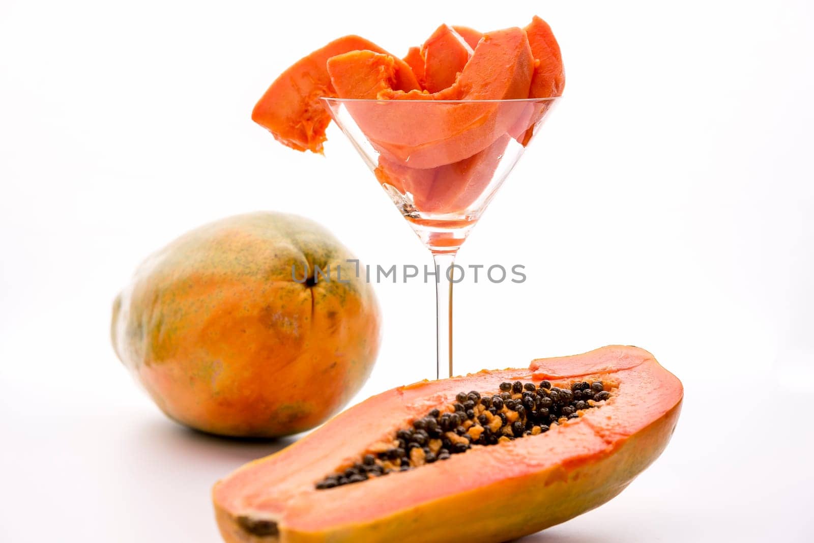 Blazing tangerine slices of the papaw fruit assorted in a long-stemmed glass. A papaw halved along its longitudinal axis in the foreground and a whole fruit in the back..