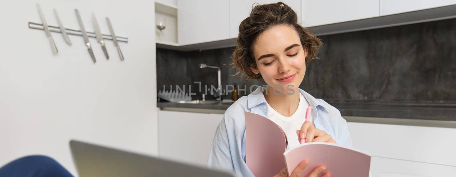 Young woman checking her schedule while working on remote from home, using laptop, looking at her daily planner, making notes, writing down information in notebook.