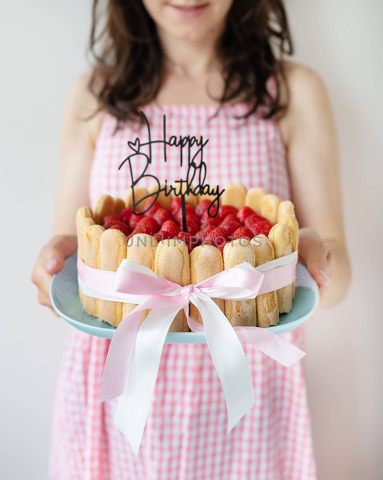 A beautiful girl in a pink dress holds in her hands a strawberry tiramisu decorated with a bow and a topper with the inscription happy birthday.