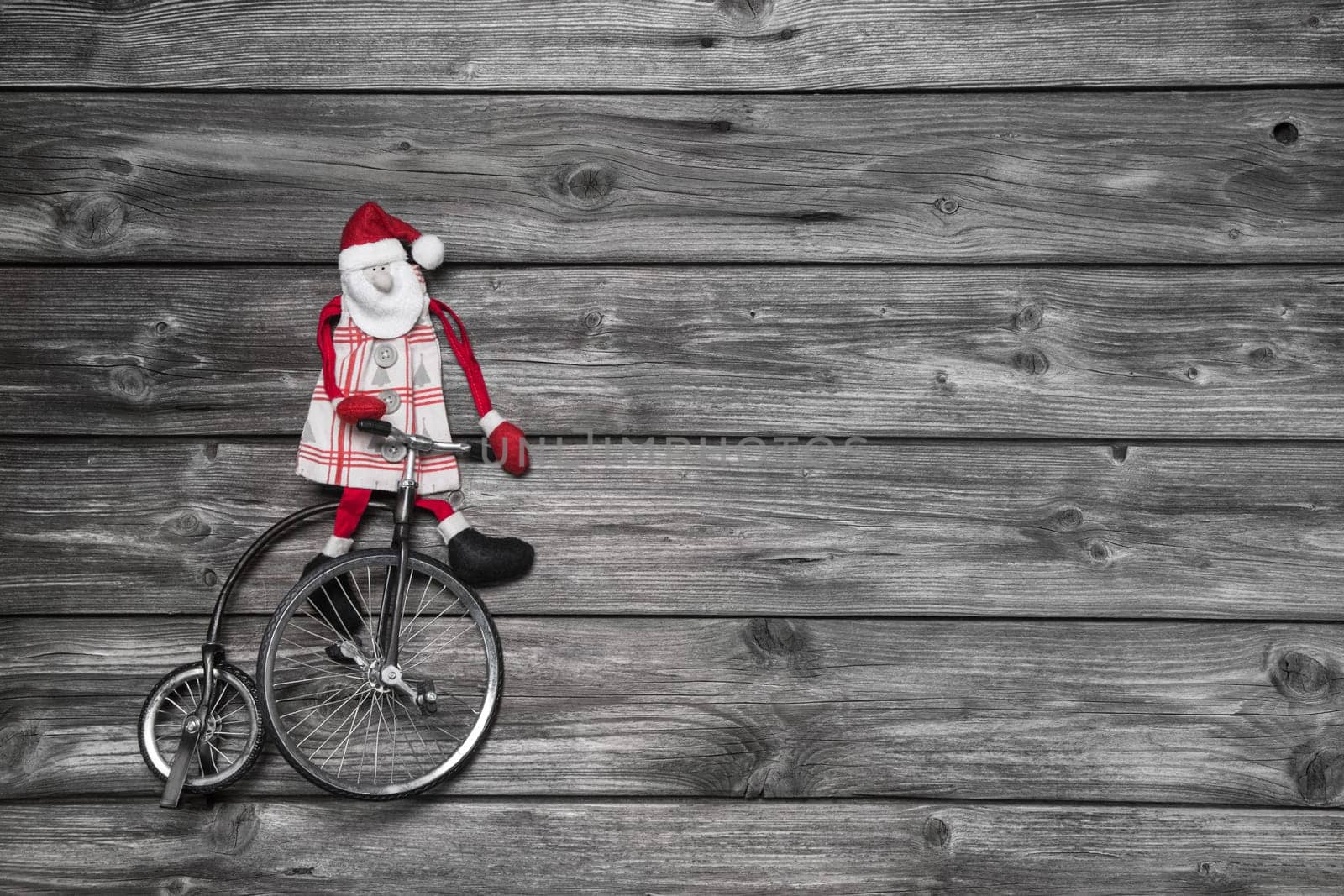 Funny red santa claus on wooden grey background in hurry for buying christmas presents sitting on a old bike.