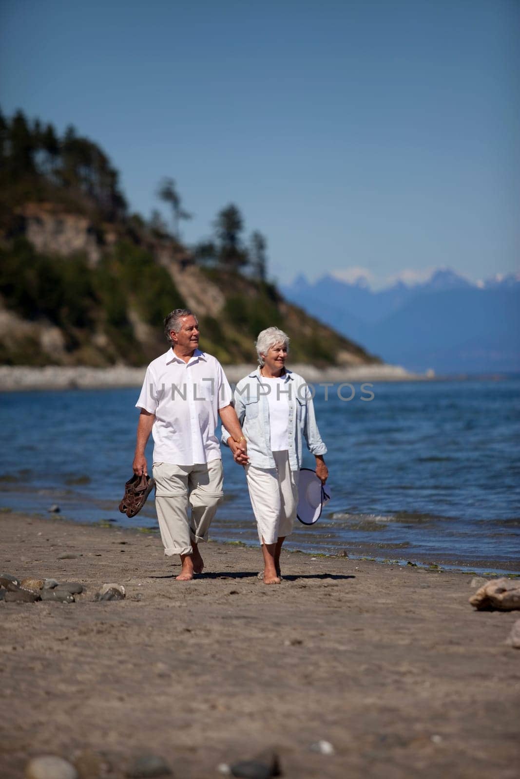 Vertical, outdoor photo of mature couple, holding hands, walking on ocean beach.
