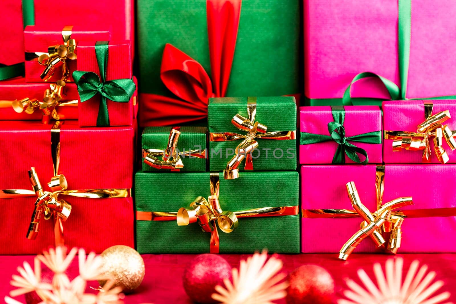Three heaps of wrapped gifts sorted by color. Red, green and magenta. All plain-colored with monochrome ribbons and bows. Tightly-framed, shallow DOF. Christmas bulbs and stars on red cloth in front.