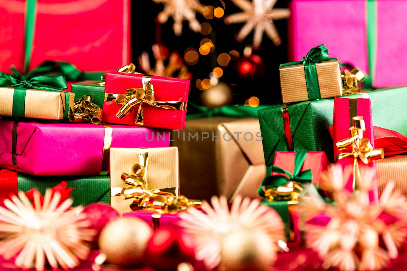 Plenty of small Christmas presents piled up before larger wrapped gifts in the rear. Narrow depth of field, tightly framed. Bokeh over black background. Blurred stars and baubles in front and rear.