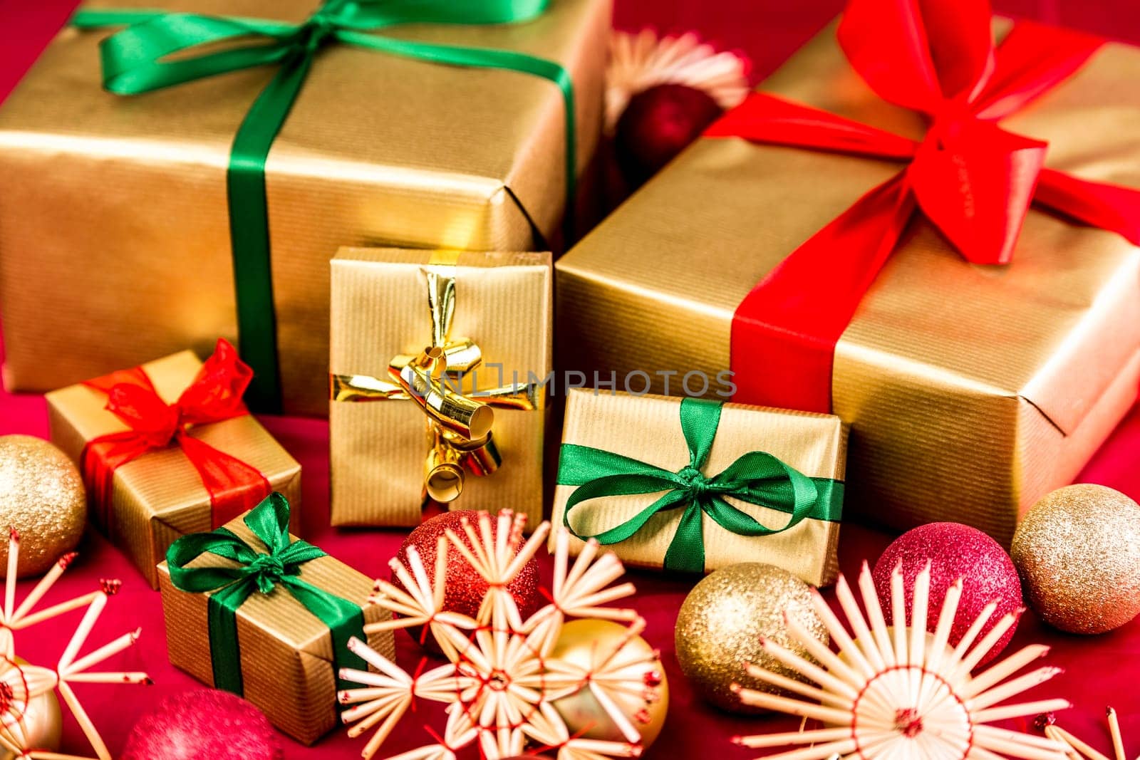 Six Christmas gifts all wrapped in gold. Bows in red and green also. Placed next to straw stars and ornaments on a red cloth. Shallow depth of field with focus on wide emerald bow around golden box.