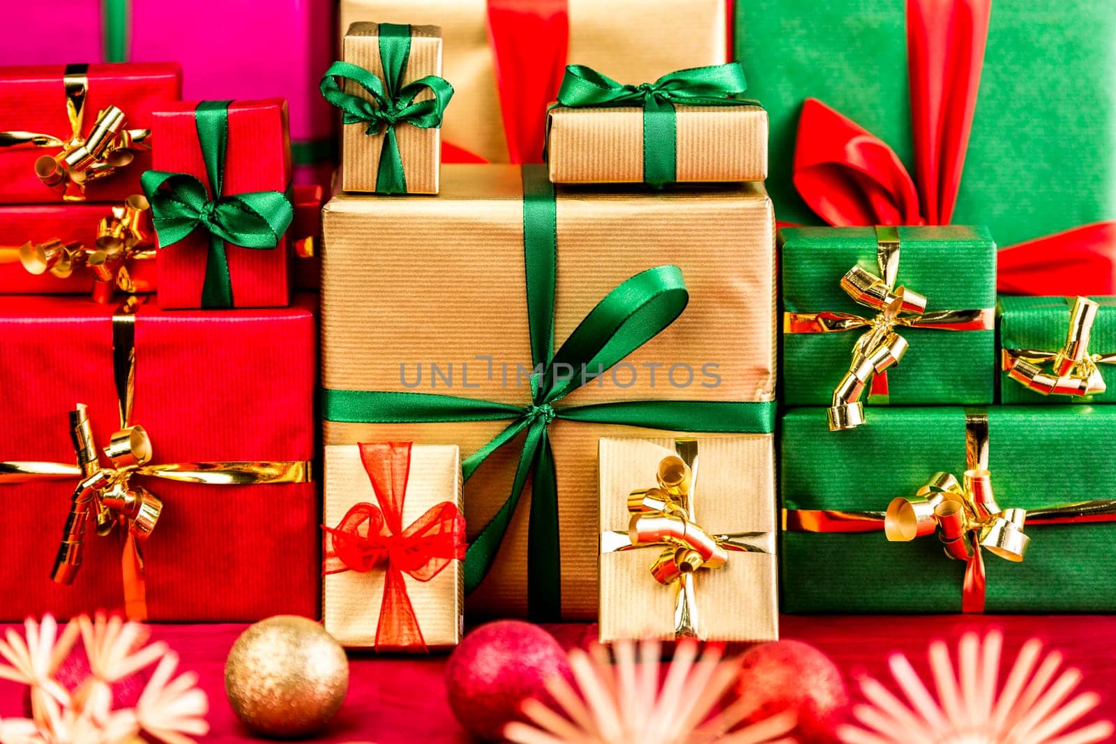 Three stacks of plain Christmas presents assorted by color. Red, gold and green. Blurred baubles and stars in foreground. Focus set on the green bow around the big golden box. Narrow depth of field.
