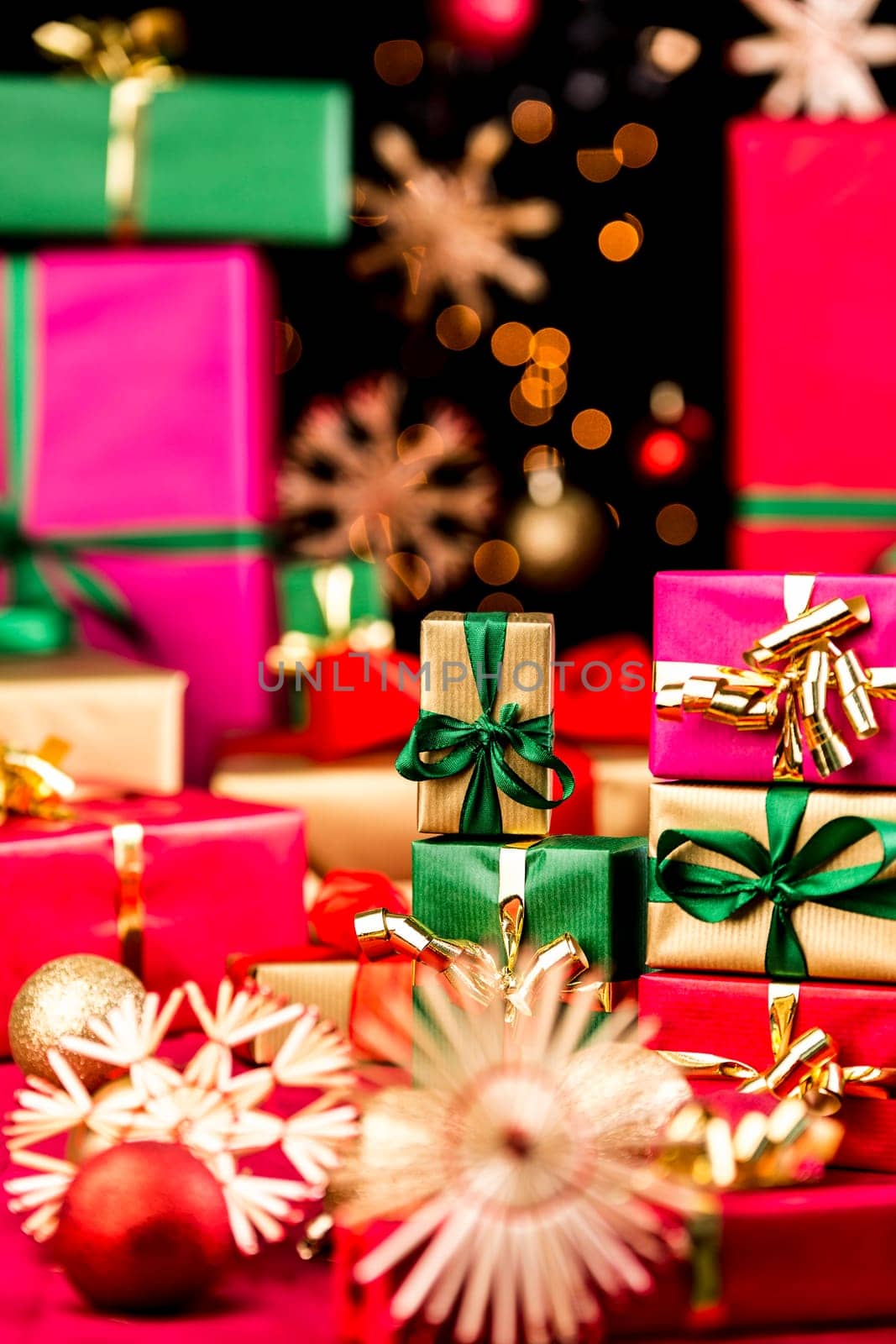 Small, single-colored Christmas presents piled up between stars and baubles. Narrow depth of field. Focus on small golden box with green bow. Out-of-focus points of light against black background.