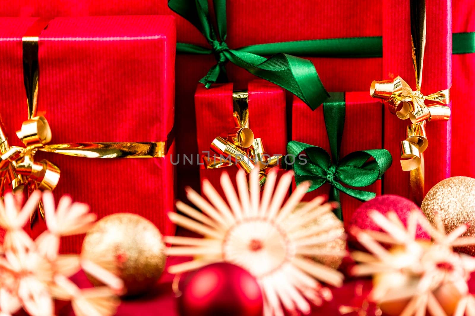 Christmas presents wrapped all in red. Green and golden bows. Blurred stars and baubles in front. Tightly framed. Flattened space. Narrow depth of field. Focus on emerald bow around smallest red box.