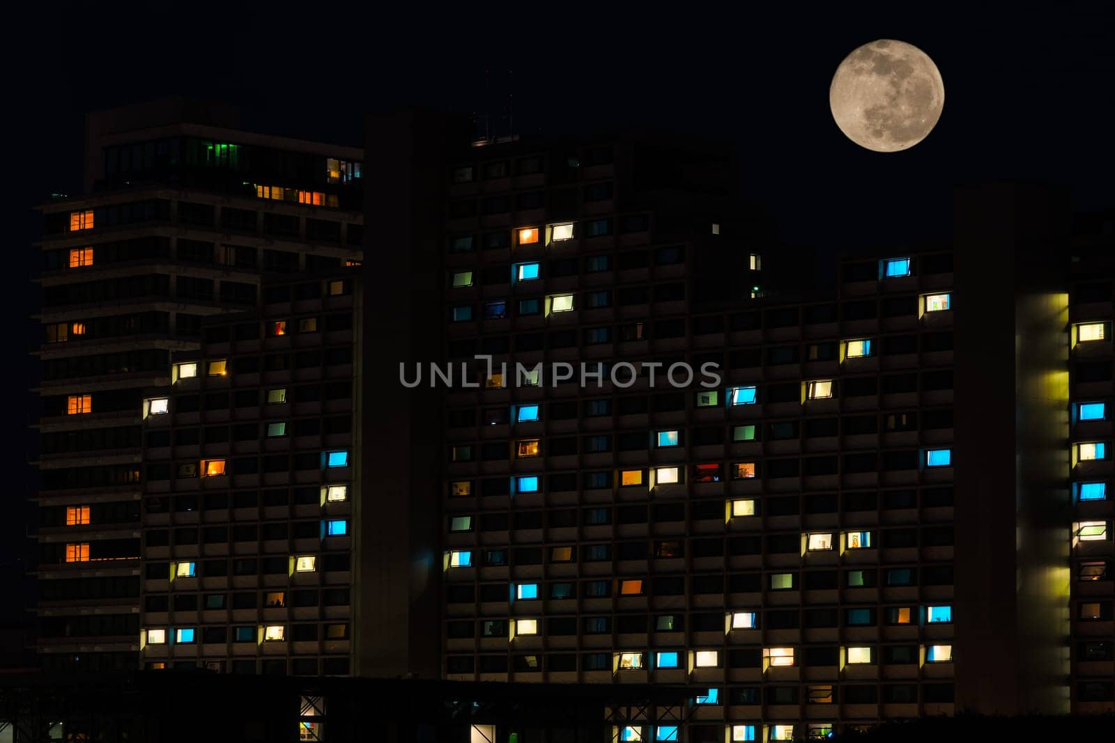 Full moon in night sky over colorful illuminated windows of residential high rise apartments house