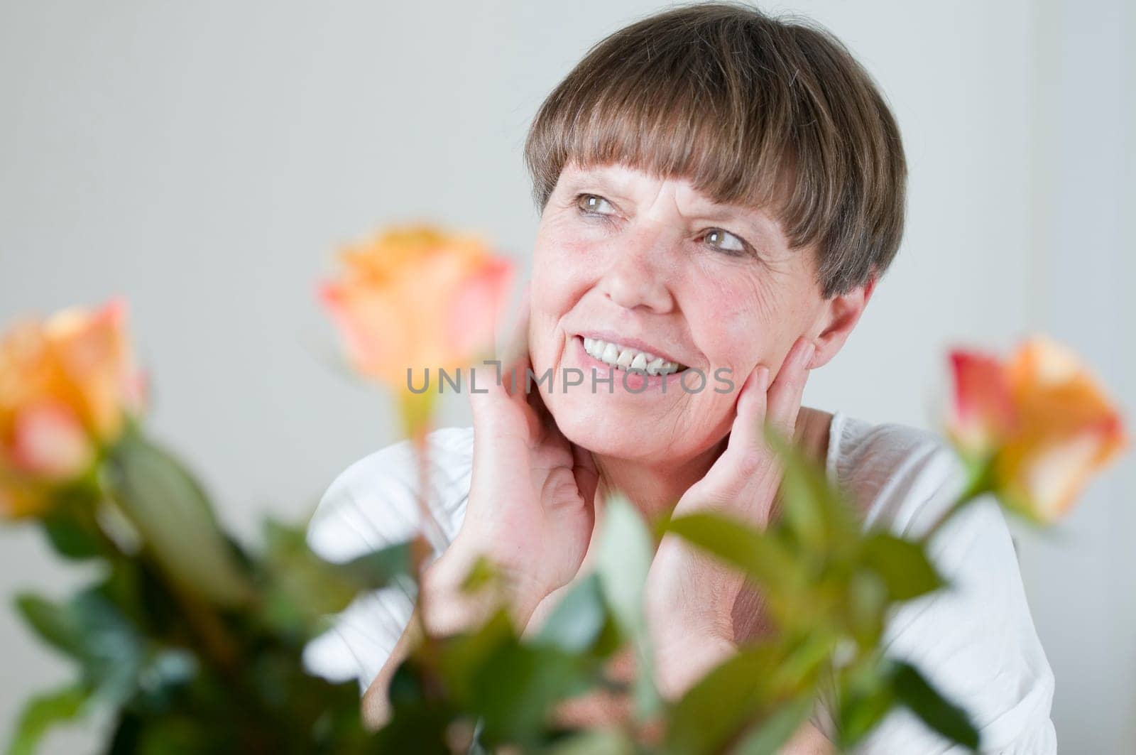 Frontal portrait of an elderly woman sitting behind flowers at the table holding her hands to her cheeks and looking obliquely up left