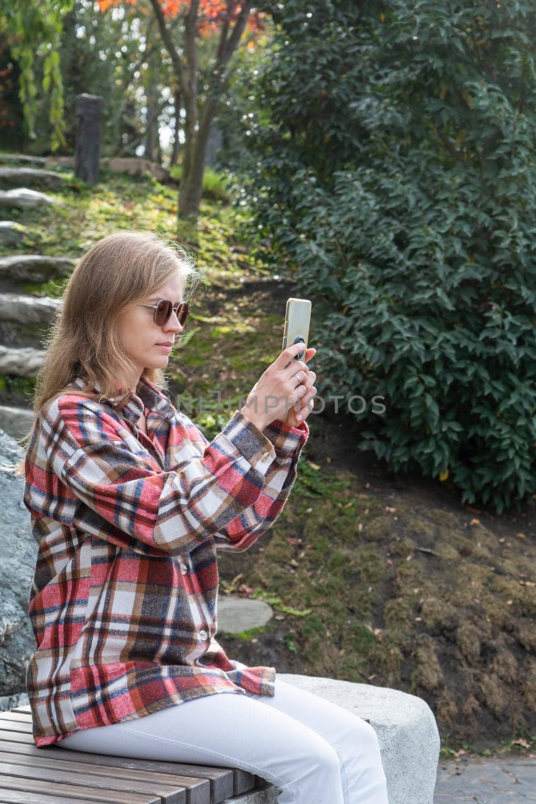 Woman in red plaid shirt enjoying nature standing in Japanese Garden taking photo on smartphone