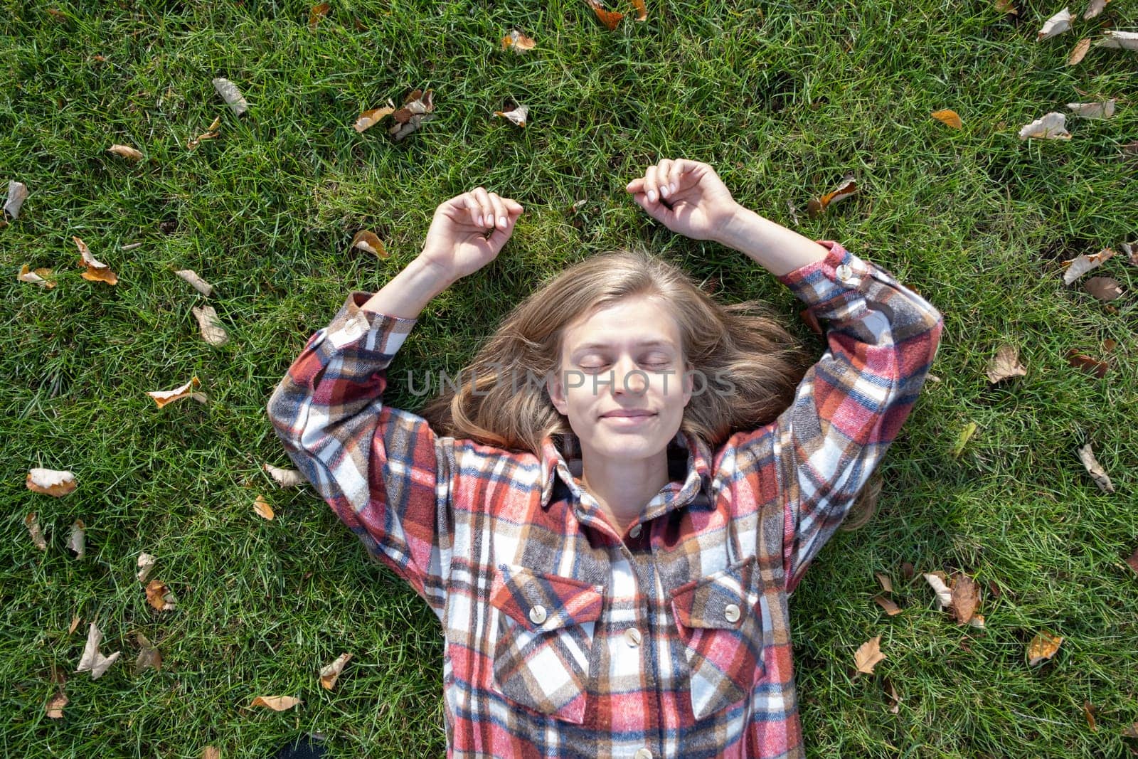 young caucasian woman in red plaid shirt smiling while relaxing on green grass with yellow autumn leaves