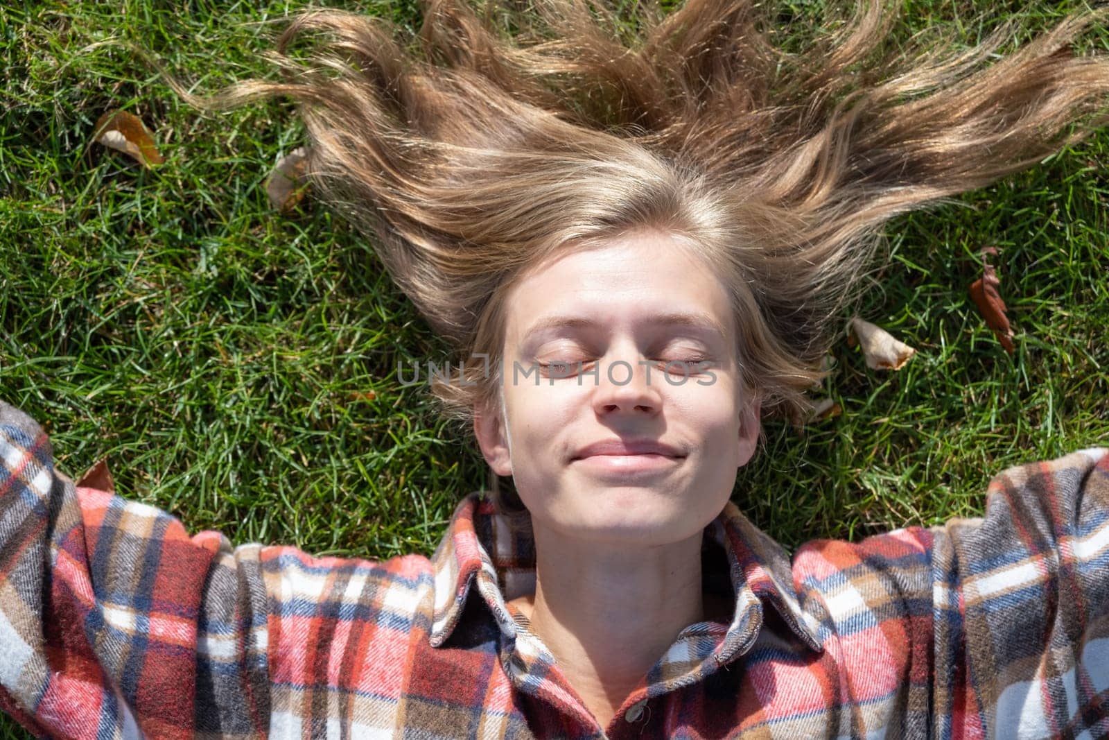 young caucasian woman in red plaid shirt smiling while relaxing on green grass with yellow autumn leaves