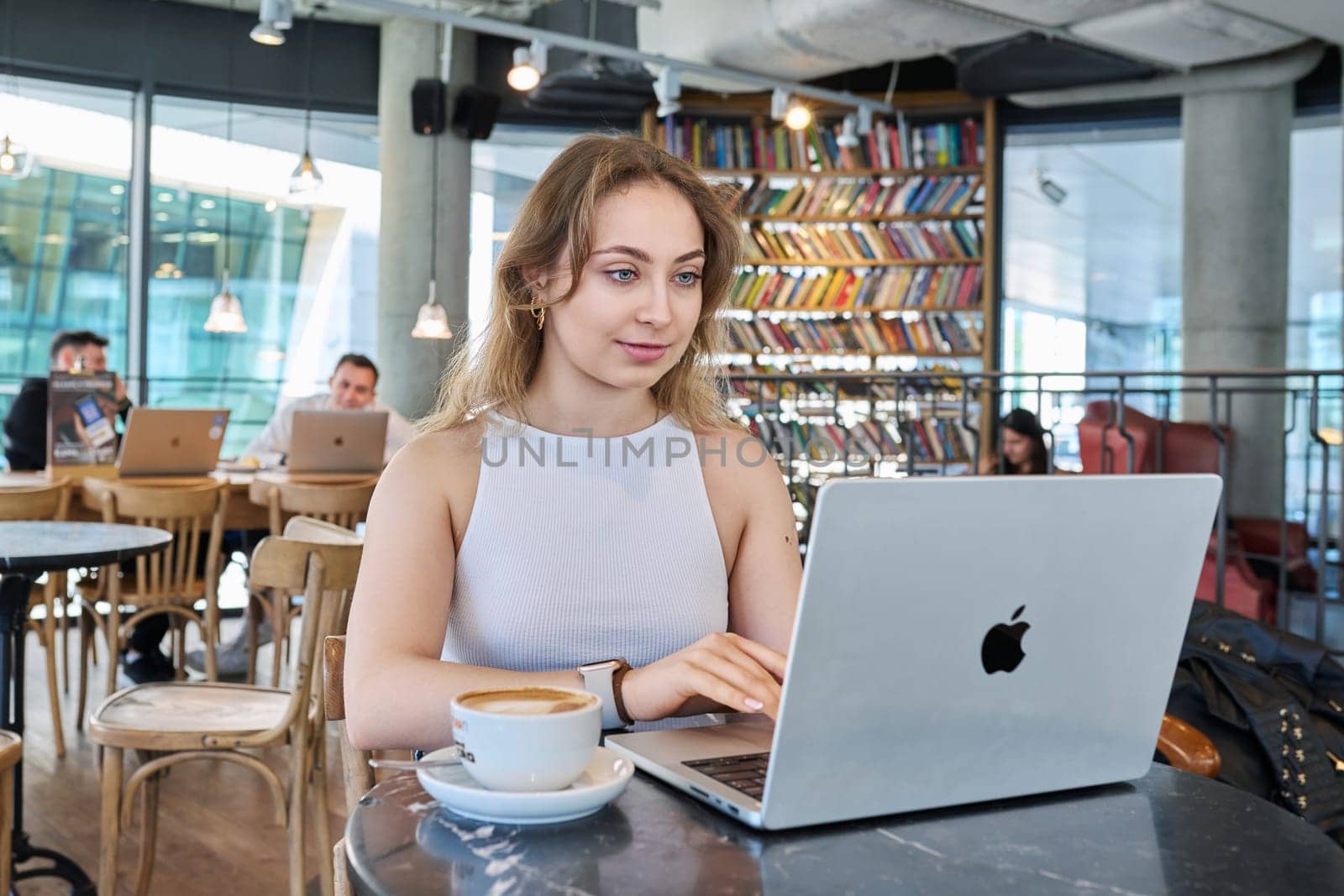 Warsaw, Poland, 12.05.2023. Beautiful girl inside cafe with cup of coffee using laptop. Attractive young blonde woman with curly hair working studying in cafe. Beauty, youth, lifestyle concept
