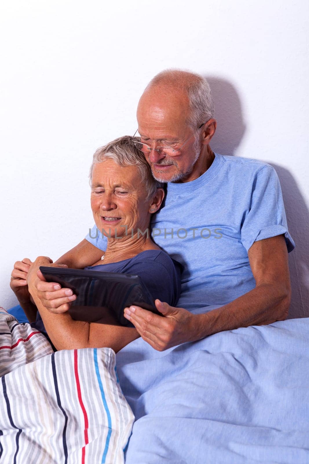 Senior couple with husband and wife lie in bed in the morning evening together with a tablet and a newspaper