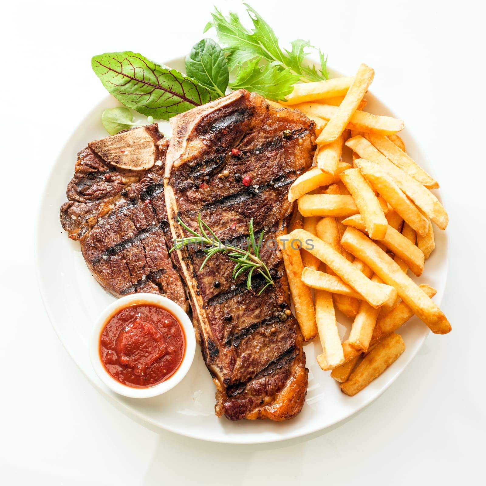 Grilled t-bone or porterhouse steak seasoned with rosemary and served with golden French fries, fresh leafy herb salad and a tomato dip, high angle view on white