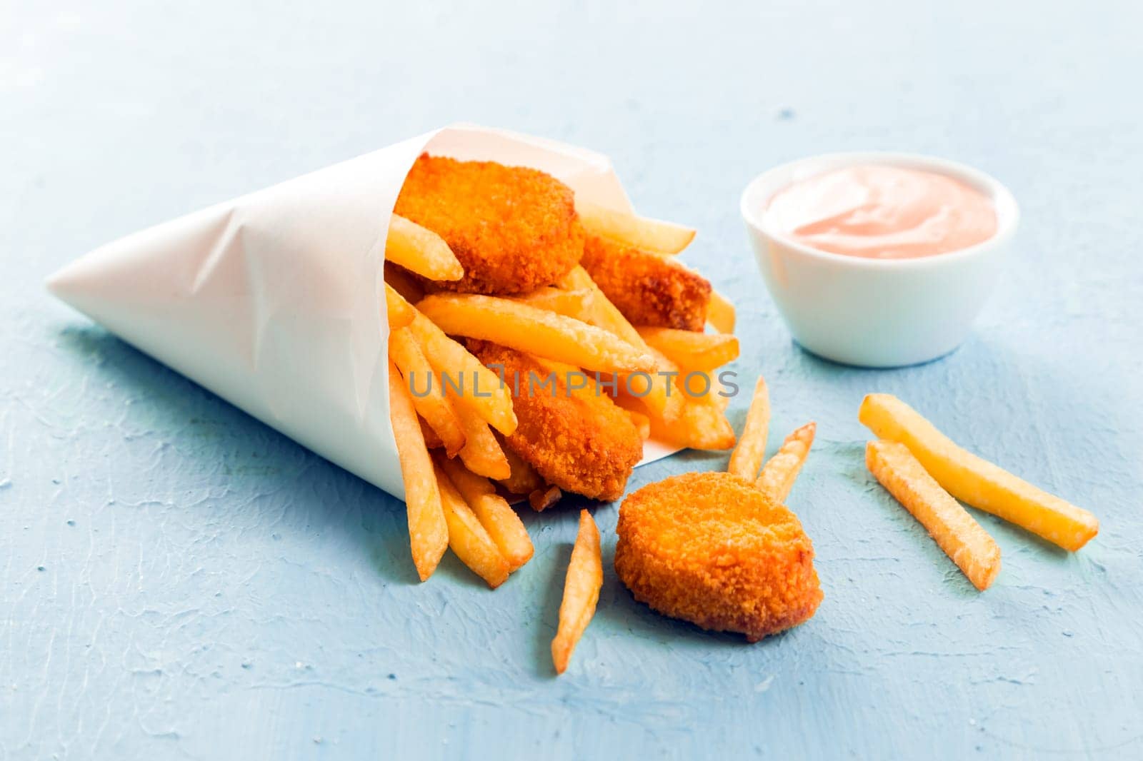 Golden fish nuggets with fried potato chips or French fries spilling out of a takeaway paper cone onto a blue surface with a small bowl of mayo dip