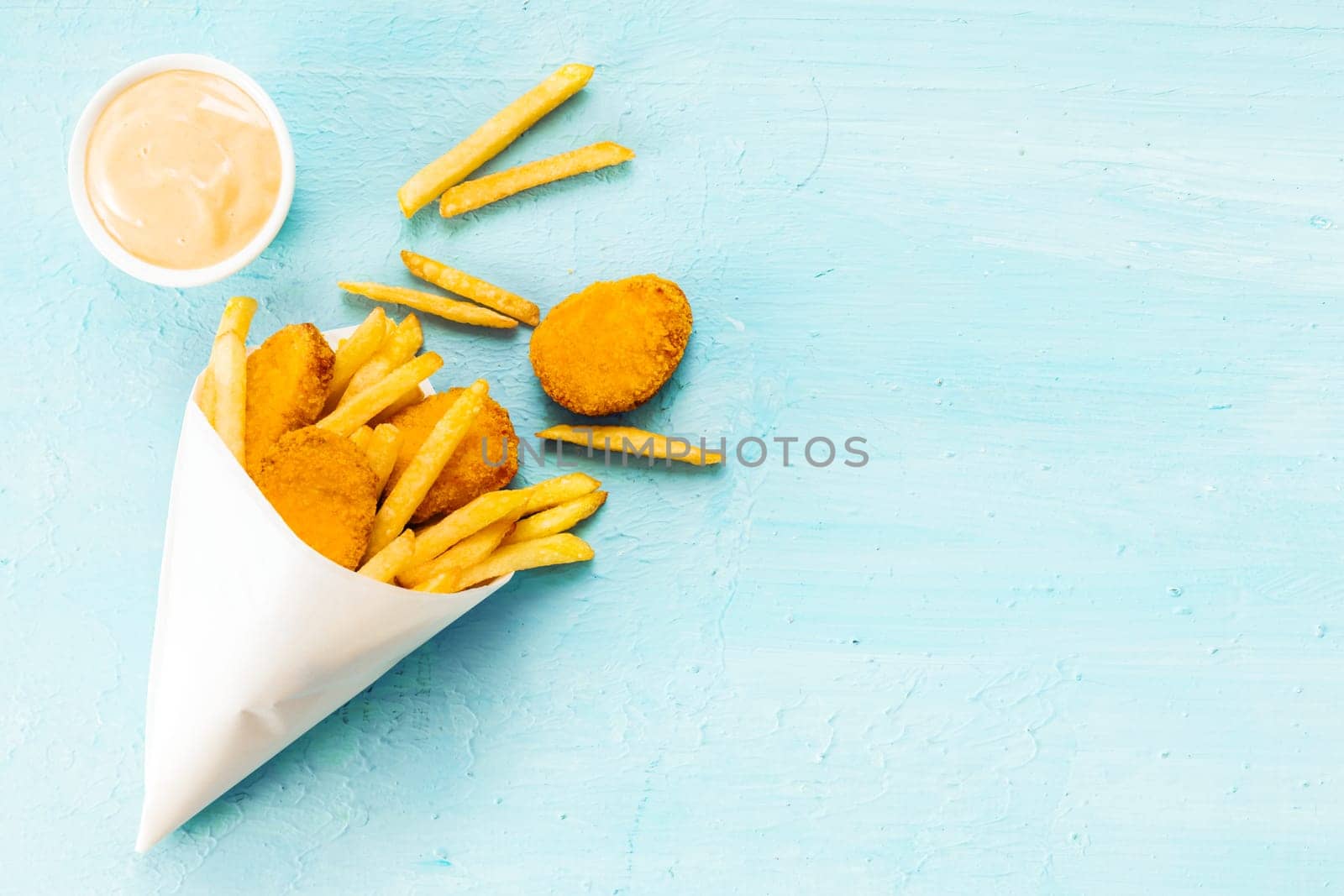 Overhead view on a blue background with copyspace of medallions of fried fish and chips in a takeaway paper cone with savory mayonnaise dip