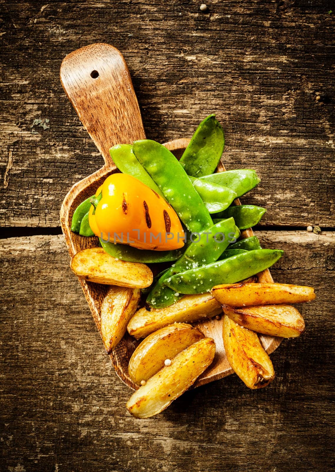 Side serving of delicious cooked vegetables served on a wooden spatula with roasted sweet red pepper, green mangetout peas and potato wedges on a rustic grunge wooden background