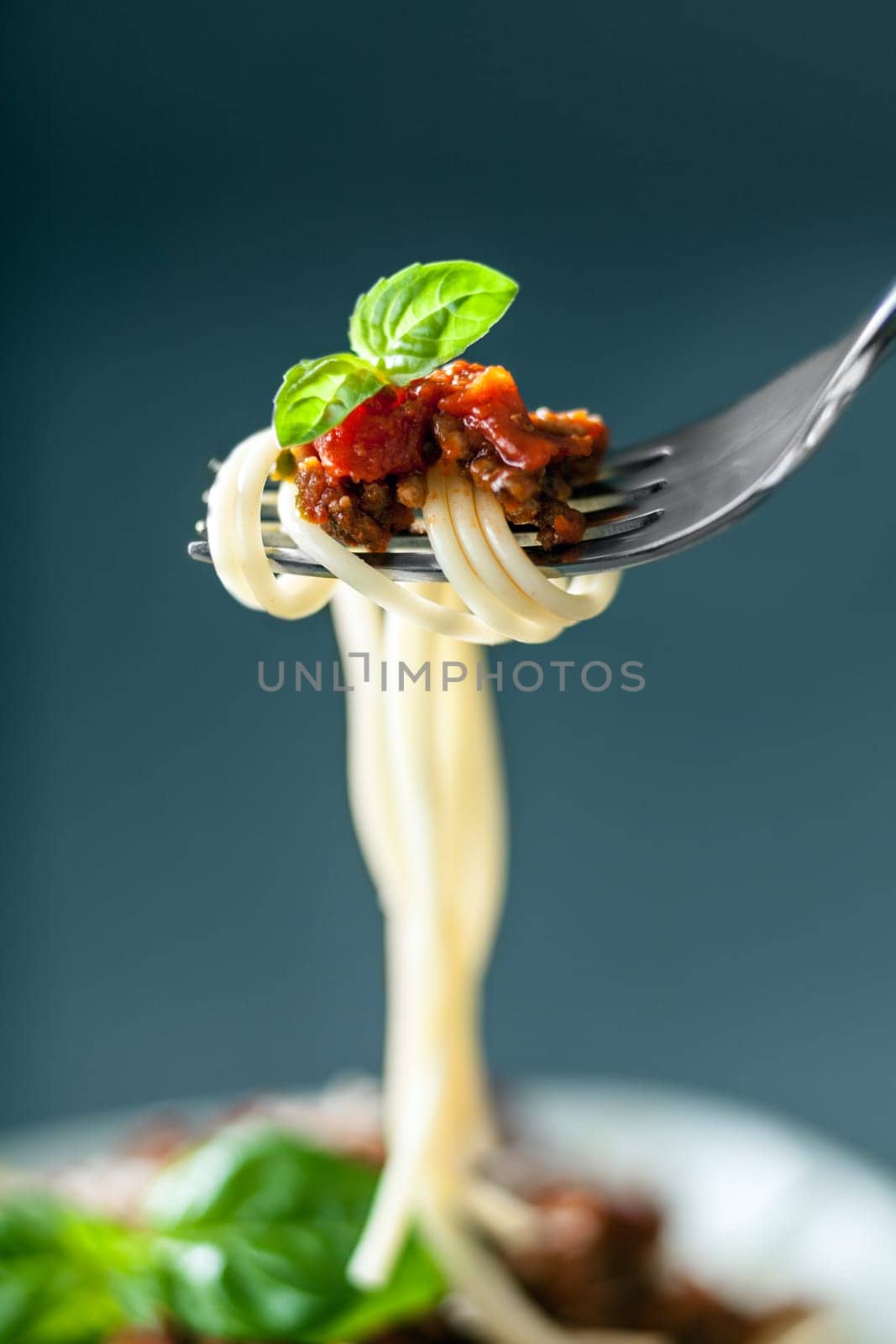 Fork full of twirled Italian spaghetti with a Bolognese meat sauce and basil suspended in the air above a full plate of food against a grey background