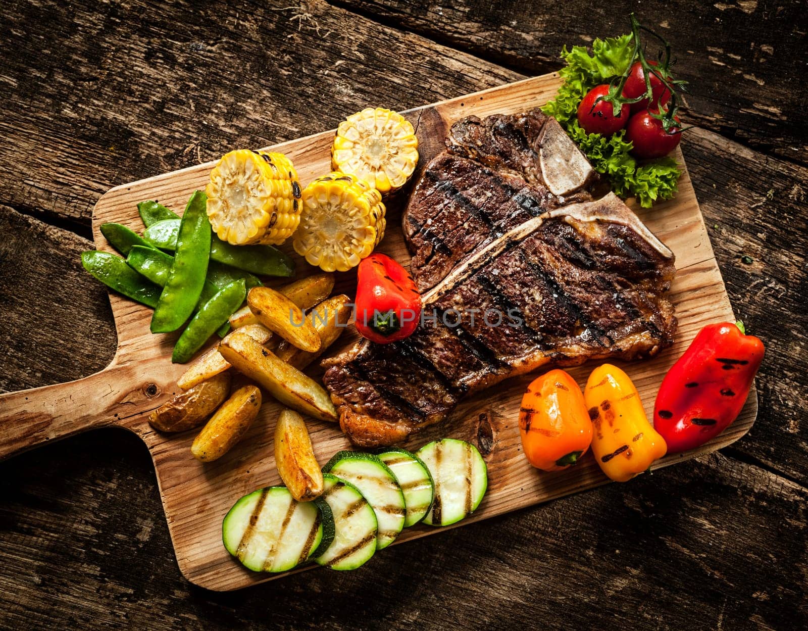 Delicious steakhouse porterhouse steak and colorful fresh roast vegetables with mangetout peas, corn, zucchini, bell pepper, potato and tomato on a wooden board, view from above