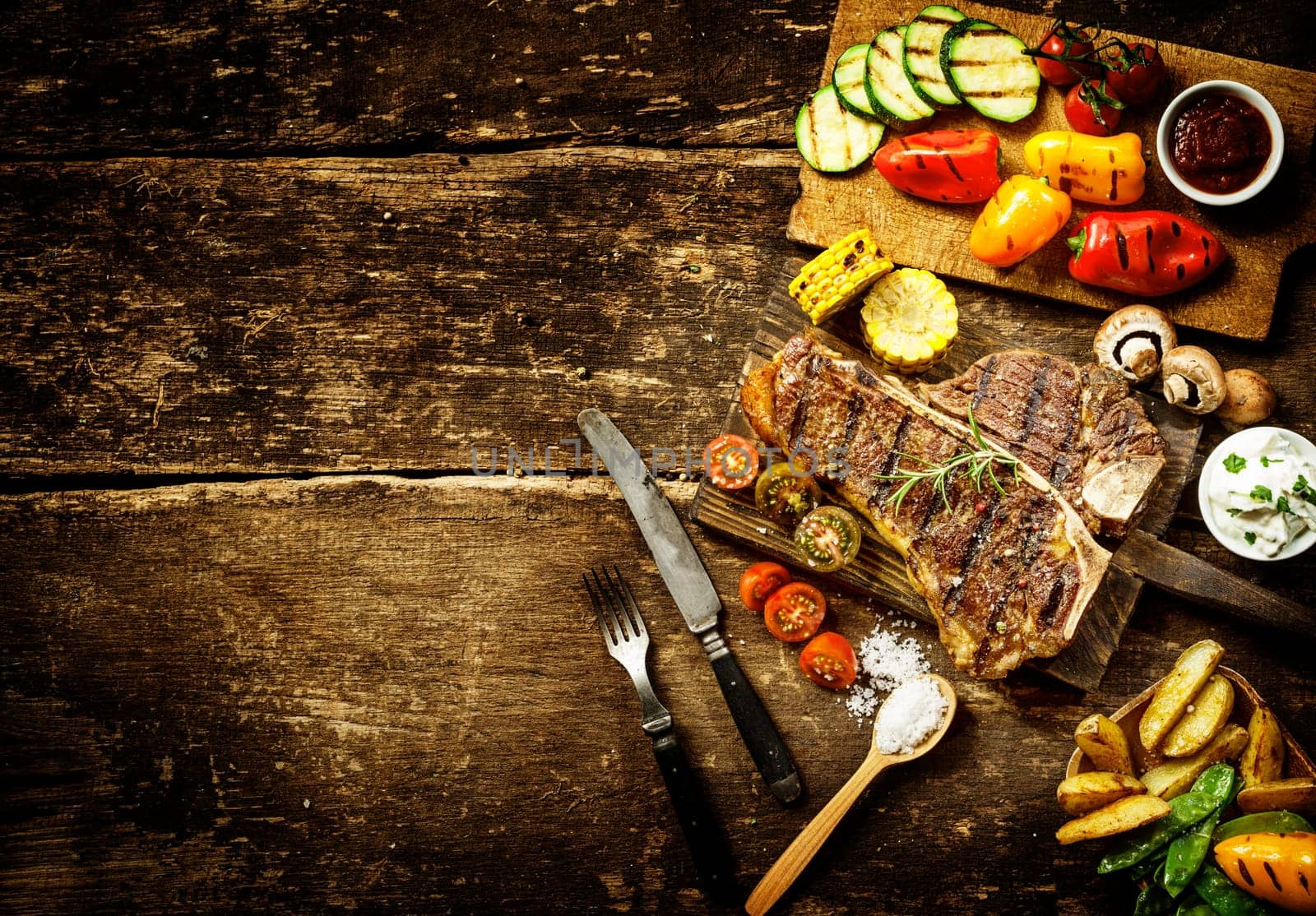 Preparing t-bone steak and roast vegetables in a country kitchen with bell peppers, mushrooms, tomato, potato, mangetout peas, and corn on a rustic wooden table with copyspace