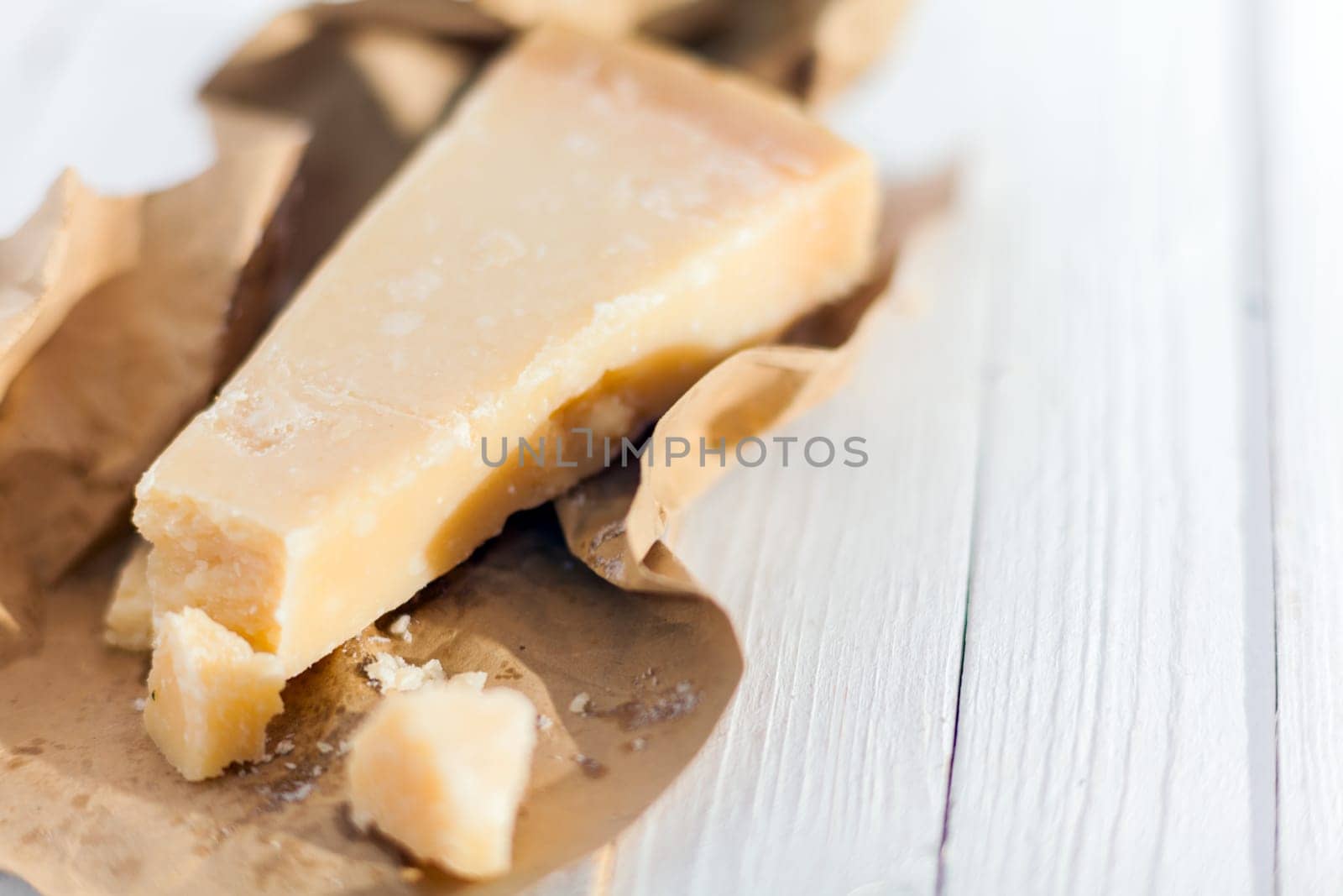 Wedge of hardy crumbly matured Italian Parmigiano-Reggiano cheese, or Parmesan, in a torn brown paper wrapper on a white wooden table