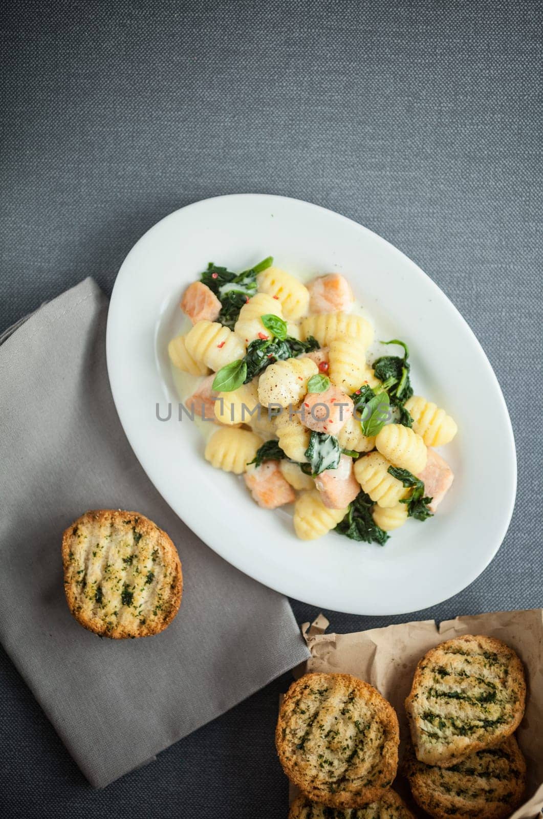 Plate of Italian gnocchi pasta and salmon pieces sauteed with fresh basil leaves and served with golden toasted crisp herbal bread, overhead view on grey with copyspace