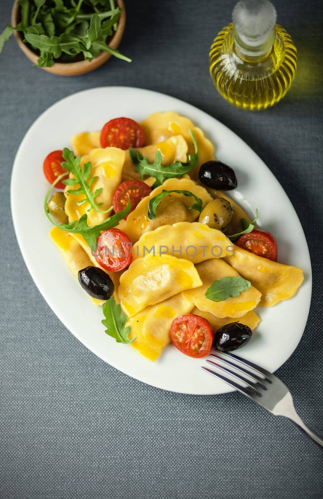 Italian ravioli pasta with olives, tomato and fresh rocket leaves served with a decanter of virgin olive oil as a dressing, overhead view on an oval platter