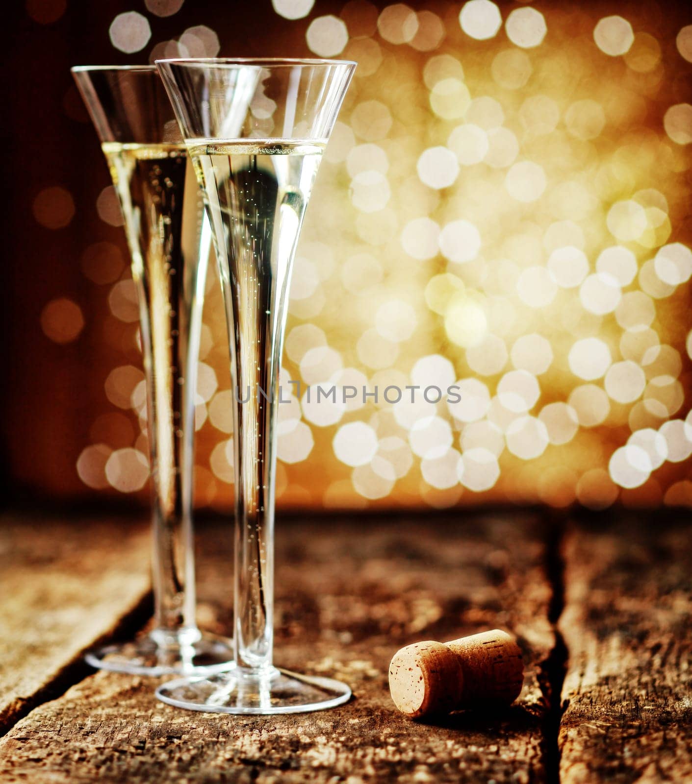 Two tall elegant flutes of romantic champagne on an old rustic wooden counter top against a sparkling bokeh of party lights celebrating a special occasion