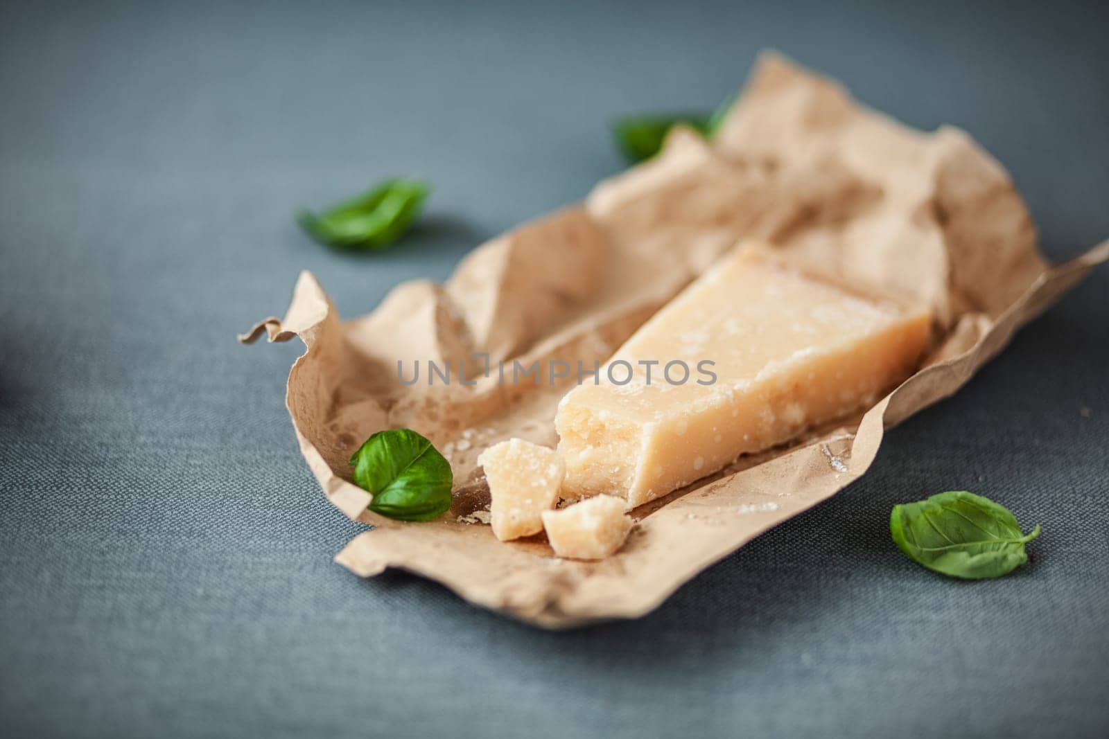 Portion of mature Italian Parmesan cheese with a traditional hard granular texture with scattered fresh basil on crinmpled brown paper, on a grey cloth with copyspace