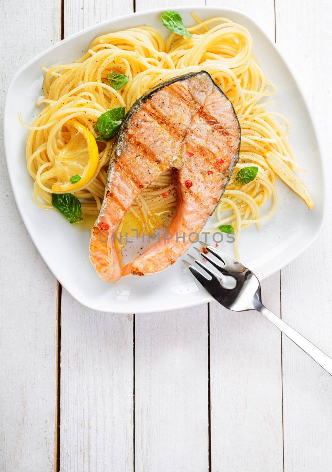Gourmet seafood cuisine with grilled salmon cutlet steak served on a bed of Italian linguine pasta garnished with lemon and fresh basil, overhead view on a white wooden table with copyspace