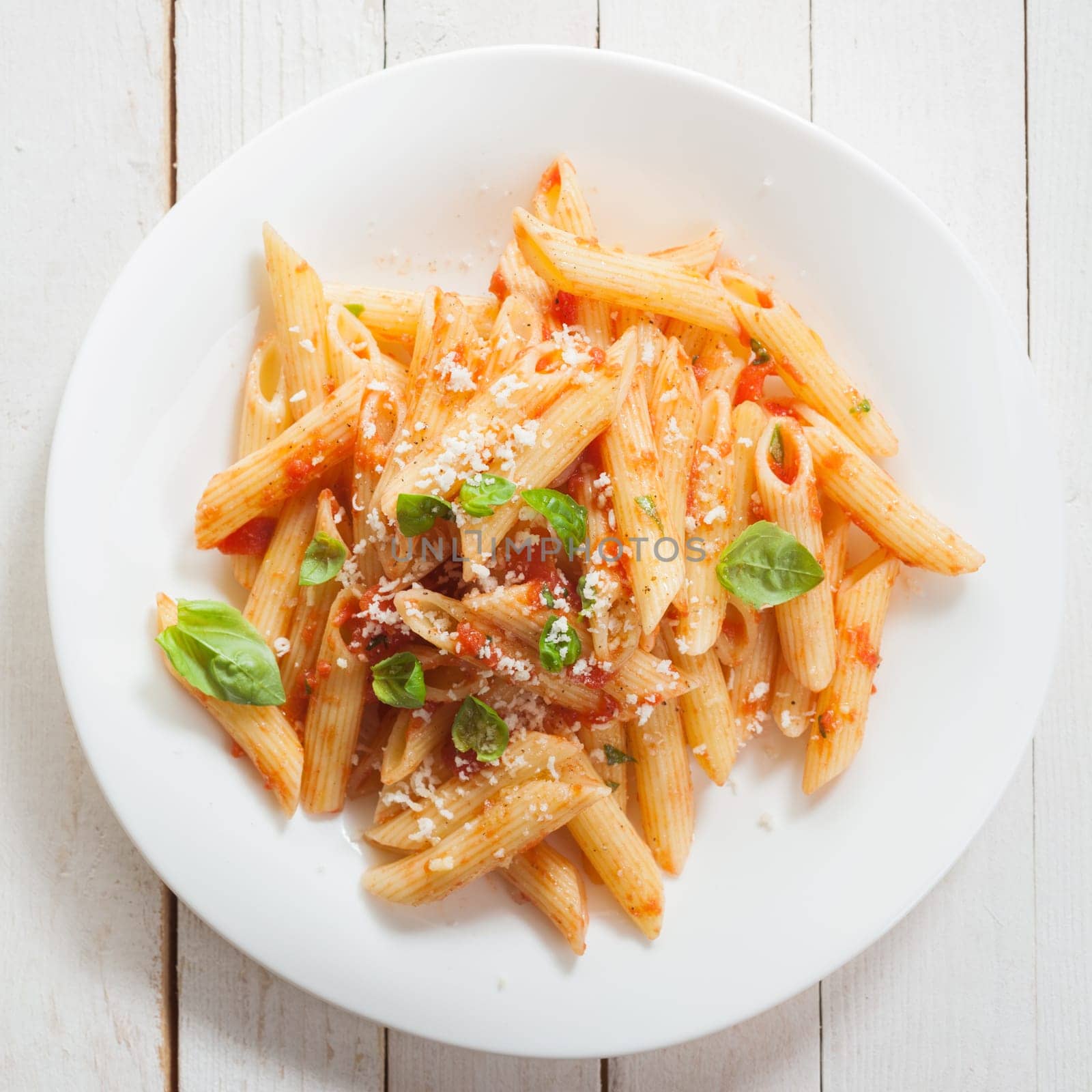 Italian penne pasta or noodles with a savory tomato sauce, fresh basil and grated parmesan cheese viewed close up from above on a white plate in square format
