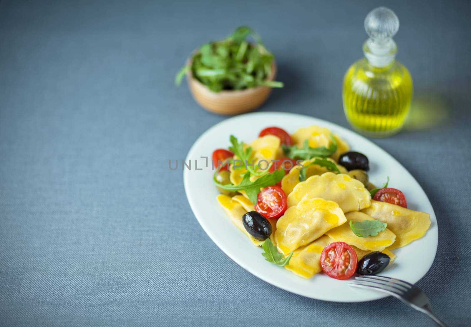 Ravioli, olive, arugula and tomato Italian salad served with fresh virgin olive oil and a side dish of fresh rocket on a grey cloth with copyspace