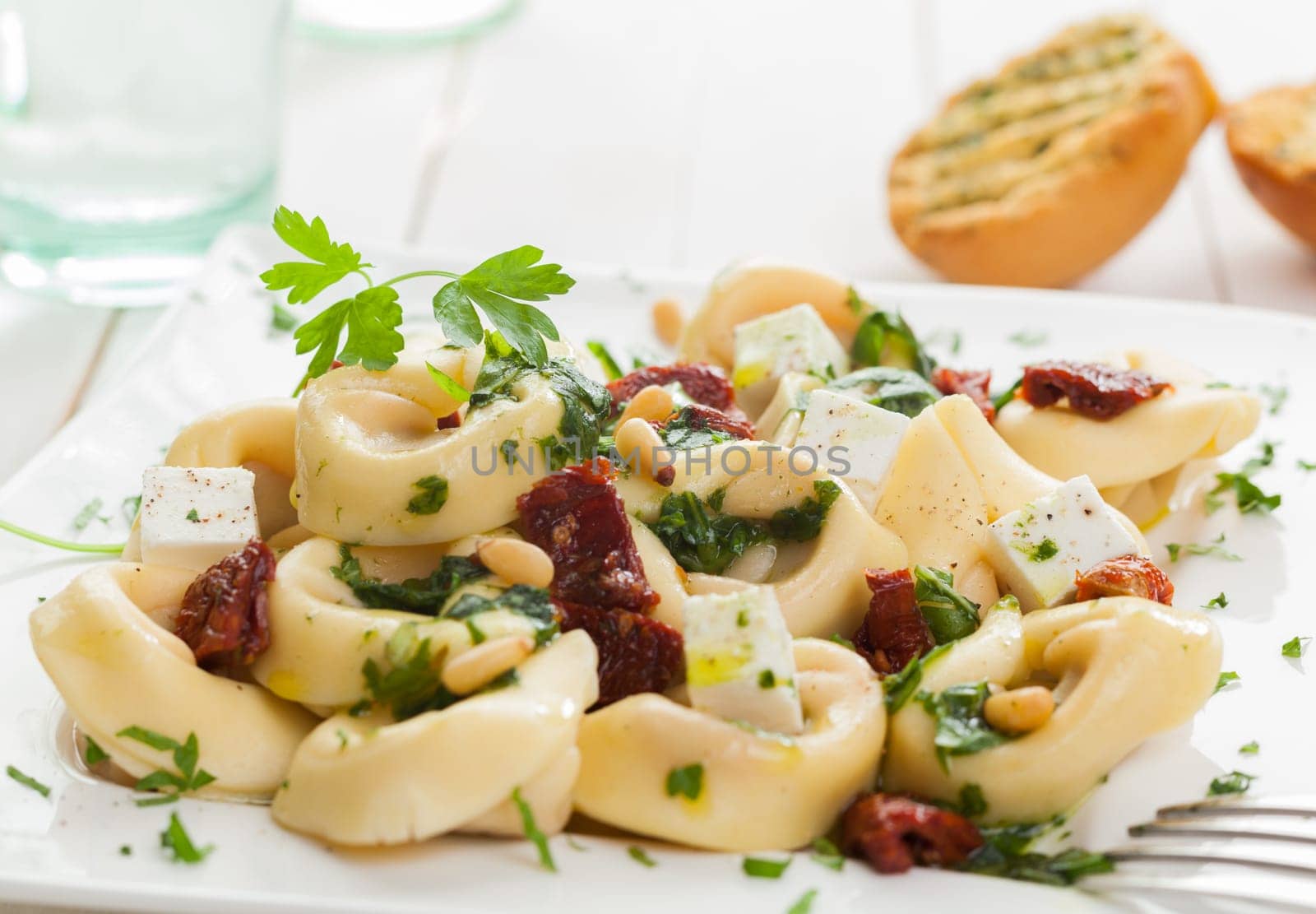 Tortellini Italian pasta topped with feta, basil, pine nuts and piquant tomatoes garnished with parsley and served with toasted grilled rolls