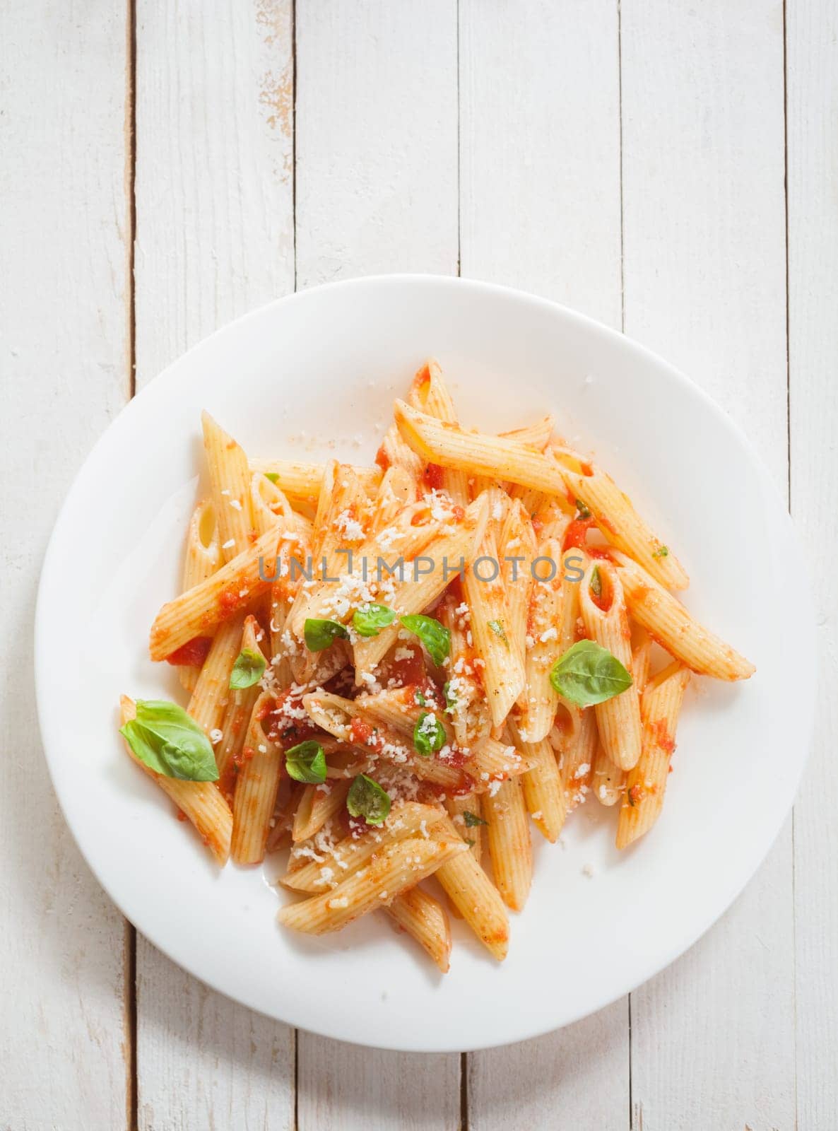 Overhead view of a healthy plate of Italian penne pasta with basil, savory spicy sauce and grated parmesan cheese on rustic white wooden boards with copyspace