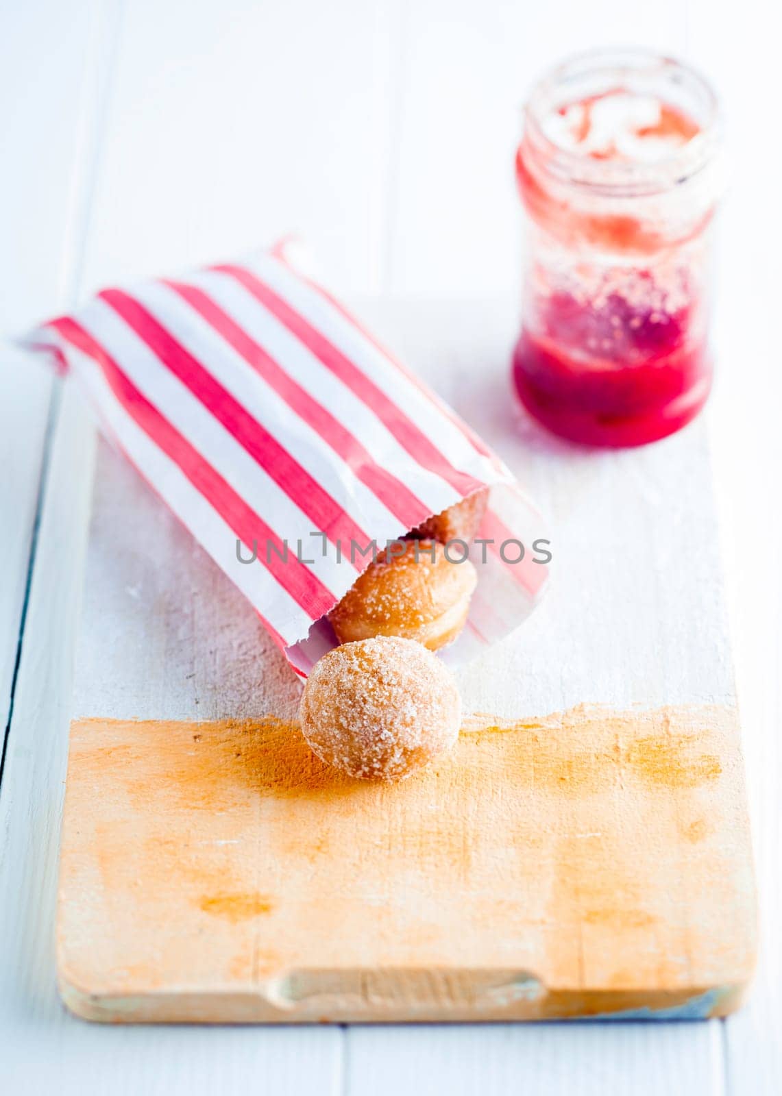 Colorful striped red and white paper takeaway packet of fresh sugared mini doughnuts on a wooden bread board with a bottle of jam or preserve behind