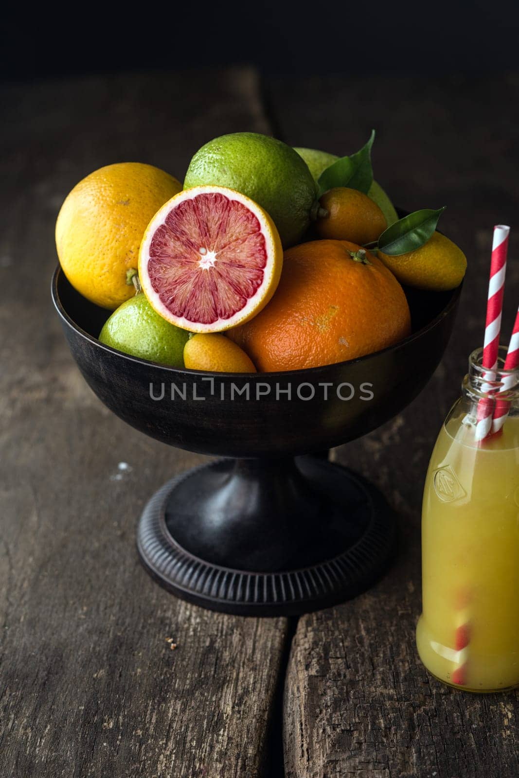 Bowl of assorted fresh citrus fruit with a halved blood orange with its distinctive red pulp in the foreground with a bottle of freshly squeezed orange juice