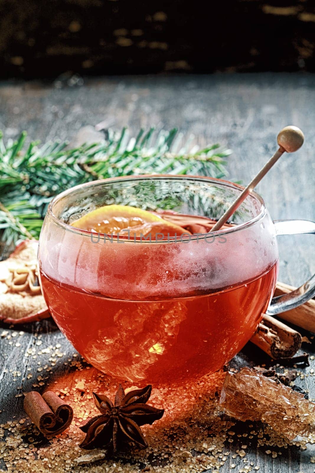 Hot cup of spiced hot tea or gluhwein with a crystallized sugar stirrer and citrus fruit surrounded by dried cinammon , star anise and caramelised sugar on a rustic Christmas backdrop with copyspace