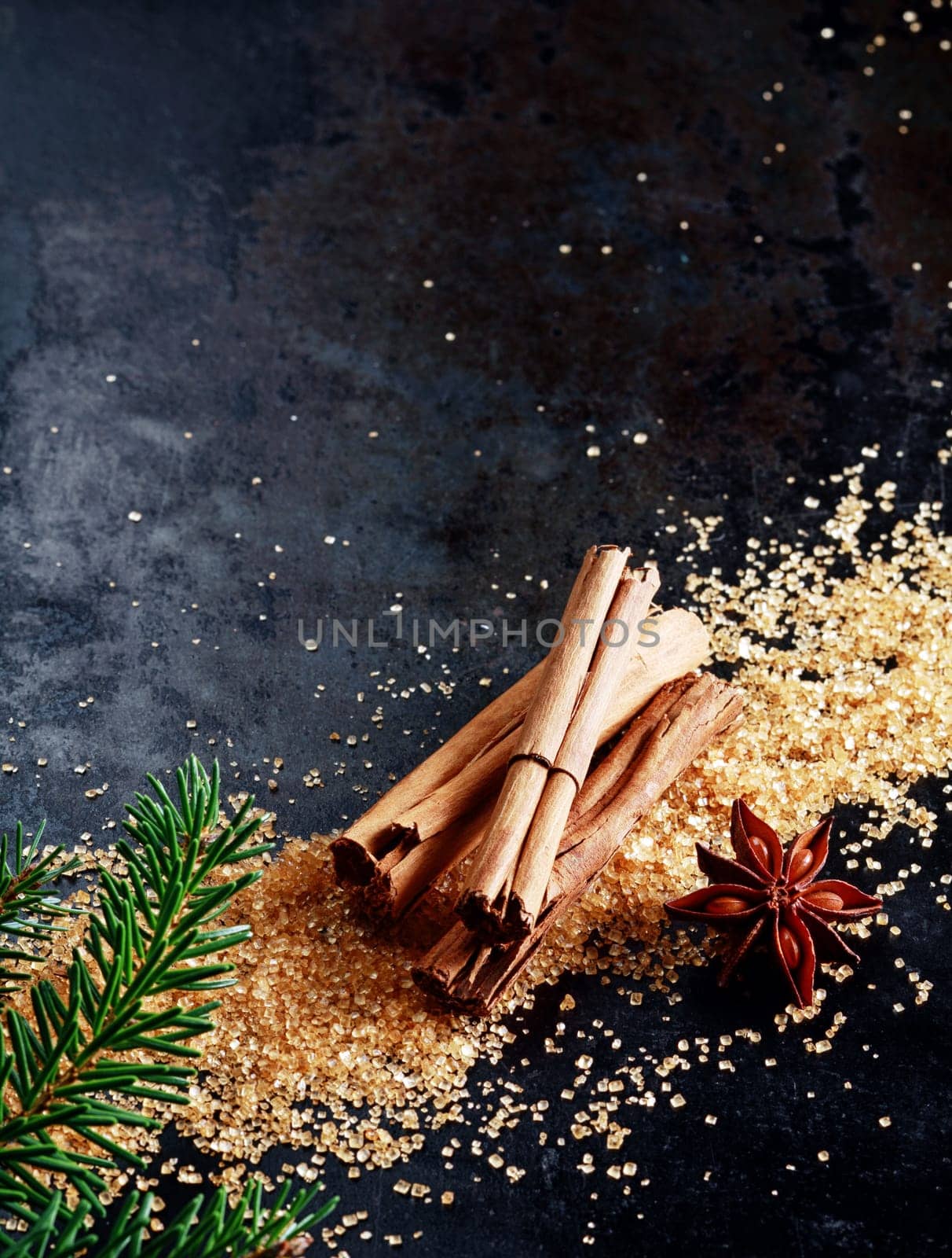 Christmas background with traditional spices with a bundle of stick cinnamon and star anise on scattered caramelized brown sugar scattered on a dark surface with copyspace and decorated with pine