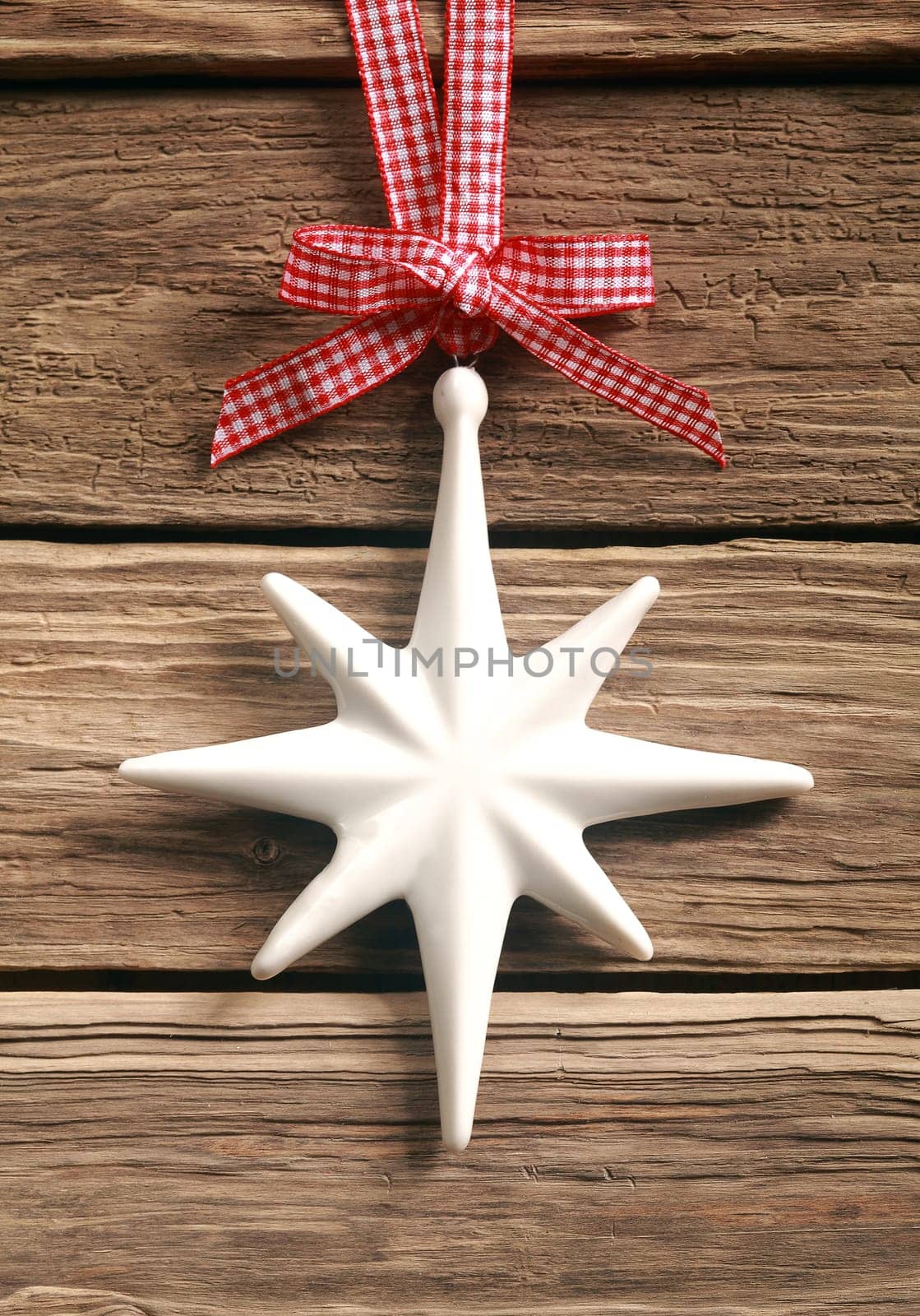 Decorative white Christmas star hanging from a rustic red and white checkered bow and ribbon on a textured weathered wooden boards background