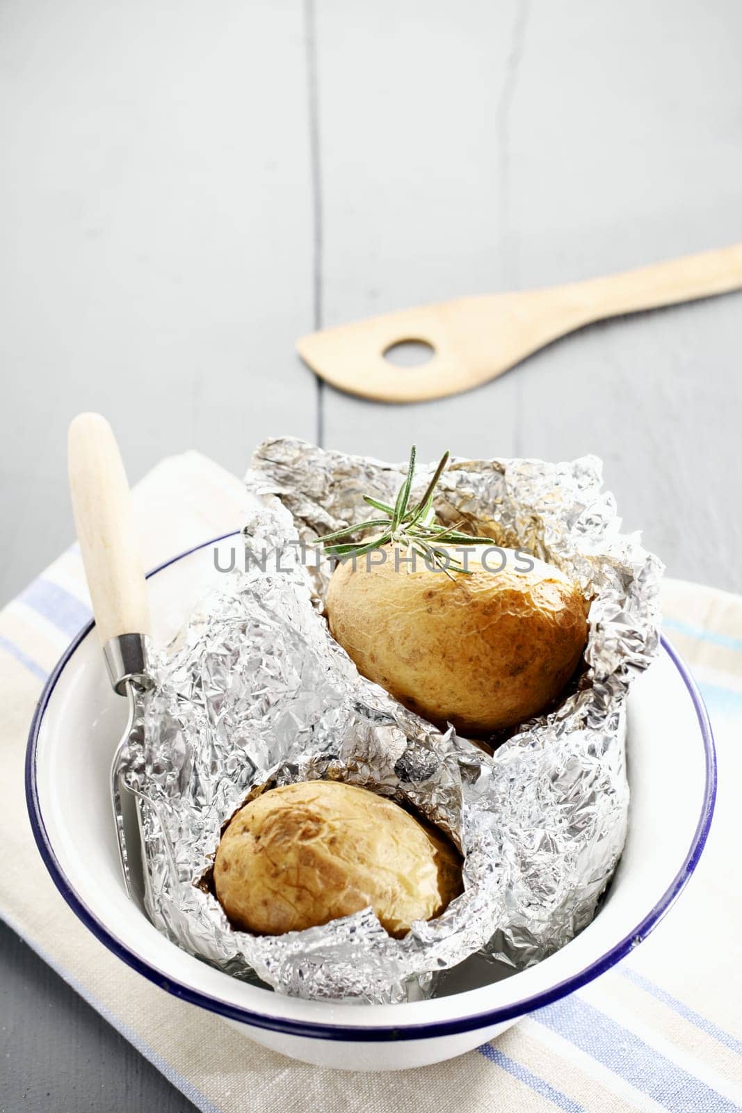 Fresh hot baked potatoes with rosemary still nestling in their foil wrappers in a metal basin on a white painted wooden picnic table, with copyspace