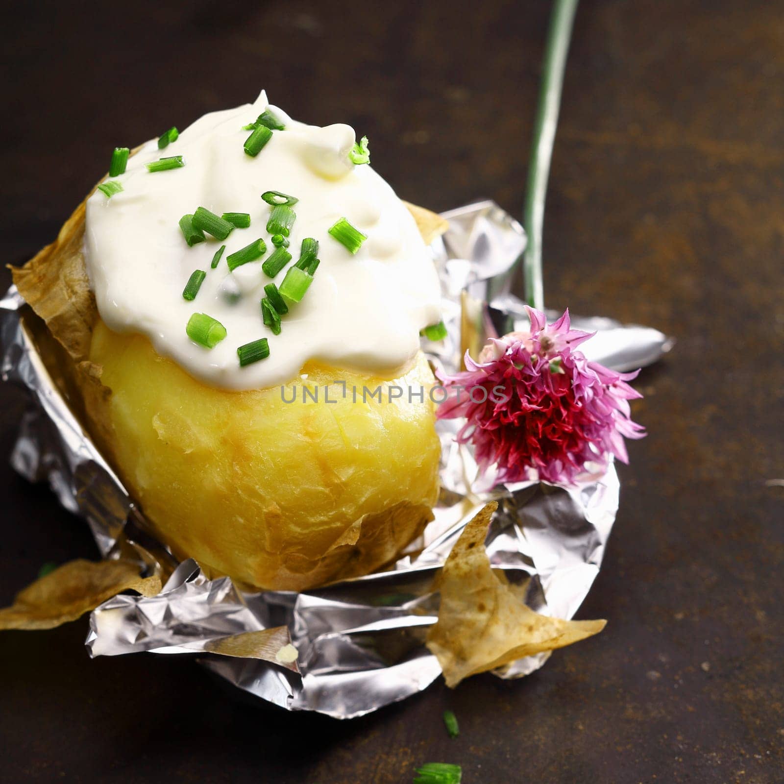 Foil baked potato with sour cream and chopped fresh chives with a flower off a chive plant served partially peeled on its foil wrapper