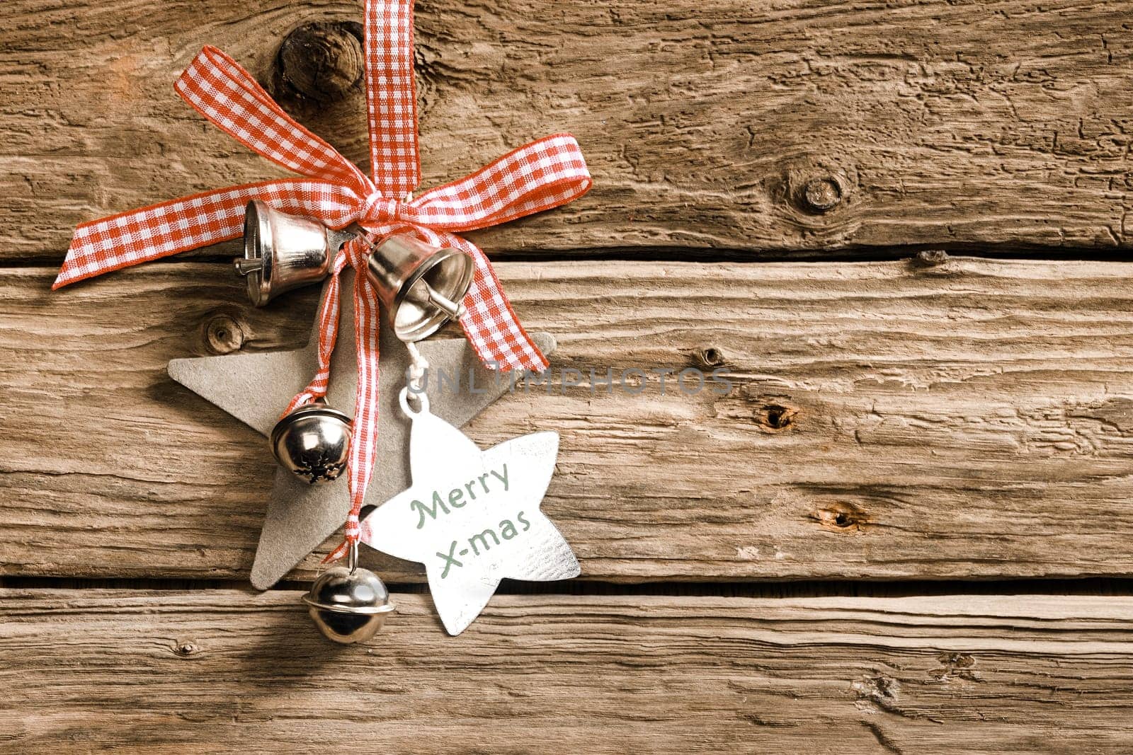 Celebrating a country Christmas with decorative stars and silver bells habging from a red and white checked ribbon and bow on rustic wooden boards with a star-shaped tag with the message - Merry X-mas